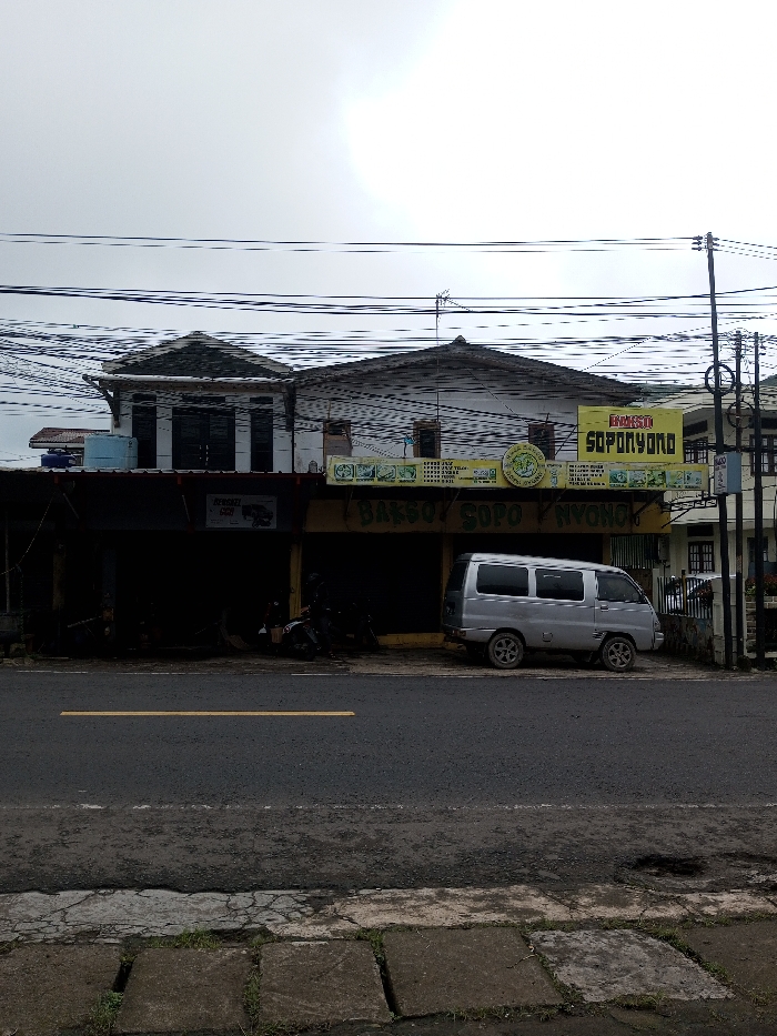 The image depicts a street-level view of a commercial building and roadway under an overcast sky. The user identified the location as the city of Neerveli, Sri Lanka; however, prominent signage on the building reads "BAKSO SOPONYONO," indicating an Indonesian business.
The scene features a two-story building in the midground, with a dark, recessed ground floor and a lighter, residential-style upper floor. Two large, rectangular yellow signs with green lettering and a central green circular logo are affixed to the facade, primarily displaying "BAKSO SOPONYONO." The ground floor consists of two dark storefront openings. A silver compact van with five windows per side is parked facing left on the shoulder in front of the right storefront. Two motorcycles are visible parked inside the dark recess of the left storefront. Overhead, numerous electrical and communication lines crisscross the frame against the bright, overcast sky. To the far right, a utility pole with additional lines and a section of another building are visible. In the foreground, an asphalt road with a single yellow center line extends horizontally, bordered by a concrete sidewalk composed of square pavers with weeds growing in the joints. No individuals are distinctly visible.