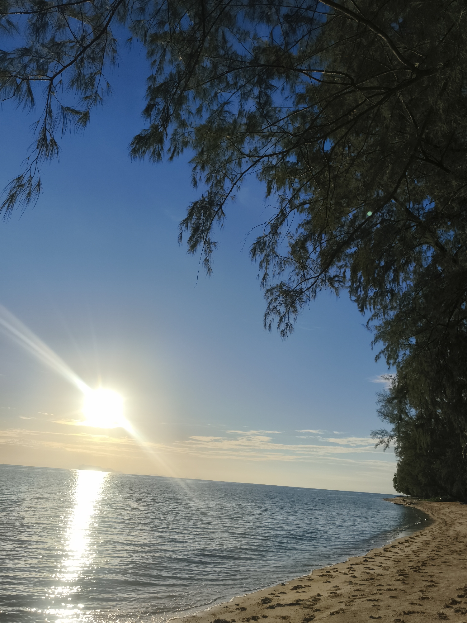 The image depicts a tranquil beach scene at Baan Lipa Noi, Thailand. The sun, positioned in the upper left quadrant, casts a brilliant reflection on the calm ocean water. The foreground features a sandy beach with visible footprints, gently meeting the lapping waves. Lush, dark green foliage from trees overhangs the top and right sides of the frame, framing the sky and sea. The sky is a clear, vibrant blue, with a few wispy clouds near the horizon. The overall impression is one of natural beauty and peacefulness, characteristic of a tropical coastal setting.