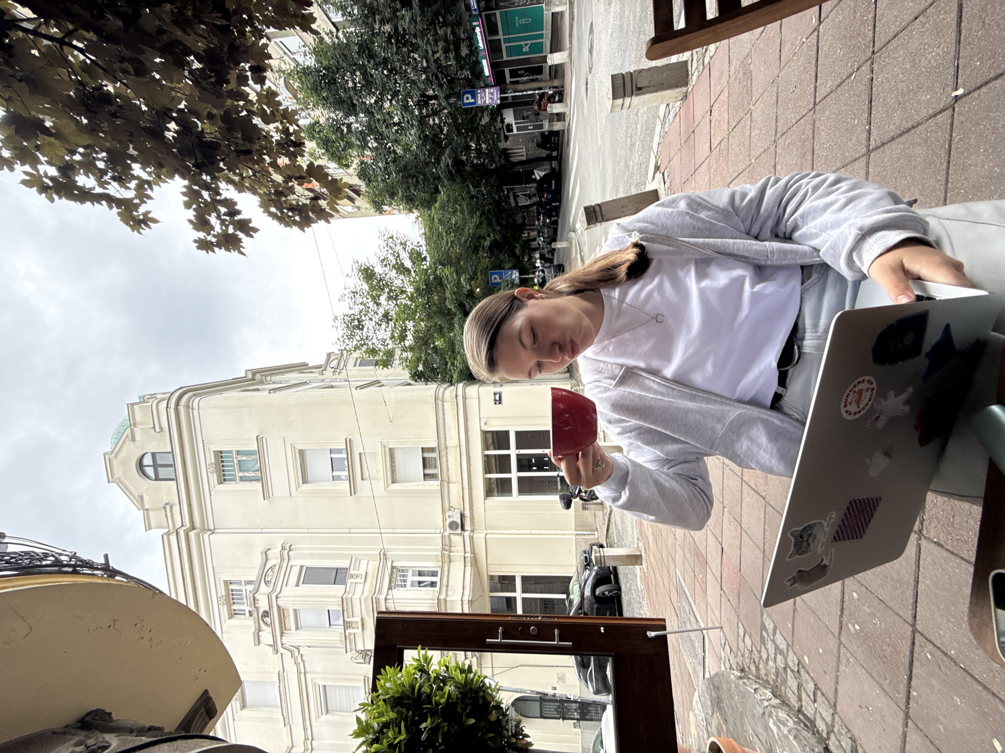 A young woman with her hair in a ponytail is sitting at an outdoor cafe table in Belgrade, Serbia, and drinking from a red mug. She is wearing a white t-shirt and a gray hooded sweatshirt, and light-colored pants. In front of her is a silver laptop decorated with stickers. The woman is looking down at the laptop, and her lips are pursed. Behind her, a street scene is visible with buildings, trees, parked cars, and traffic. The day appears to be overcast.