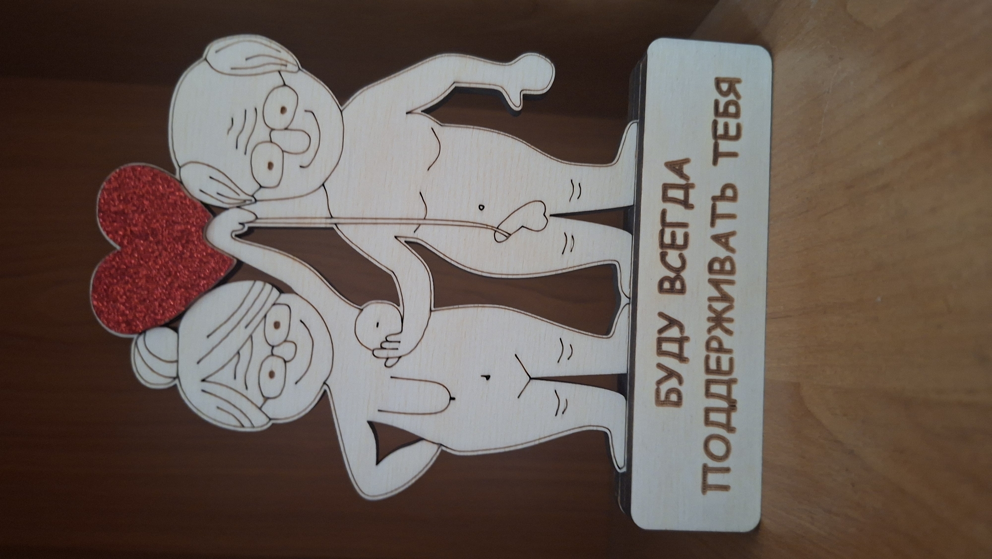 A wooden, laser-cut figurine depicts a stylized, elderly couple – a man and a woman – standing intimately on a rectangular base. The figures are light-colored wood with engraved outlines for facial features and body details, suggesting a nude or semi-nude representation with anatomical details.
The man, with glasses and a receding hairline, has a smiling or amused expression. His left arm is around the woman's shoulder or upper back, and his right hand rests on her belly. The woman, with hair in a bun and bangs, also has a smiling expression. She holds a vibrant red, glittery heart between them with both hands, and her left hand is positioned near the man's groin area, which features an engraved erect penis. Both figures show details like nipples and belly buttons. Their bodies are slightly intertwined, conveying closeness and affection.
The figurine stands on a dark brown surface, likely a table or shelf. On the right side of the wooden base, engraved in Cyrillic, is the text "БУДУ ВСЕГДА ПОДДЕРЖИВАТЬ ТЕБЯ", which translates to "I WILL ALWAYS SUPPORT YOU". The overall impression is one of enduring love, intimacy, and playful support between an elderly couple.
