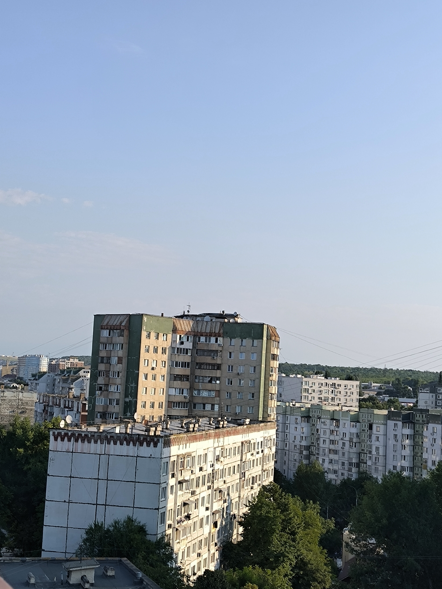 This is a high-angle, long-shot photograph of a residential area in Chișinău, Moldova, on a clear day. The sky is a pale blue, with a few faint wisps of clouds near the horizon. In the foreground, a weathered, multi-story apartment building with a white facade and a patterned concrete upper section dominates the lower right quadrant. This building exhibits numerous balconies and windows, some with air conditioning units. Behind it, and slightly to the left, is another, taller apartment building, predominantly green on its upper floors, with a rusty, corrugated metal roof structure. This building also features many windows and balconies. In the background, a panorama of similar apartment buildings stretches across the city, interspersed with lush green trees and some distant structures. Power lines are visible crisscrossing the middle ground. The overall impression is of a typical urban landscape in an Eastern European city. There are no people or animals visible in the photograph, and no specific actions or interactions are depicted.