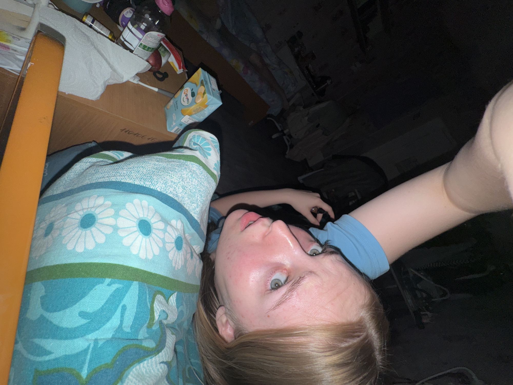 A photograph, likely a selfie, captures a pale-skinned person with blonde hair, lying on a bed covered with a blue and green patterned blanket. The person's head is near a wooden table, cluttered with various items including boxes, bottles, and a juice carton. The background is dark, suggesting a dimly lit room, with indistinct shapes indicating furniture or other objects. The location is not identifiable as Perm, Russia from the visual content.