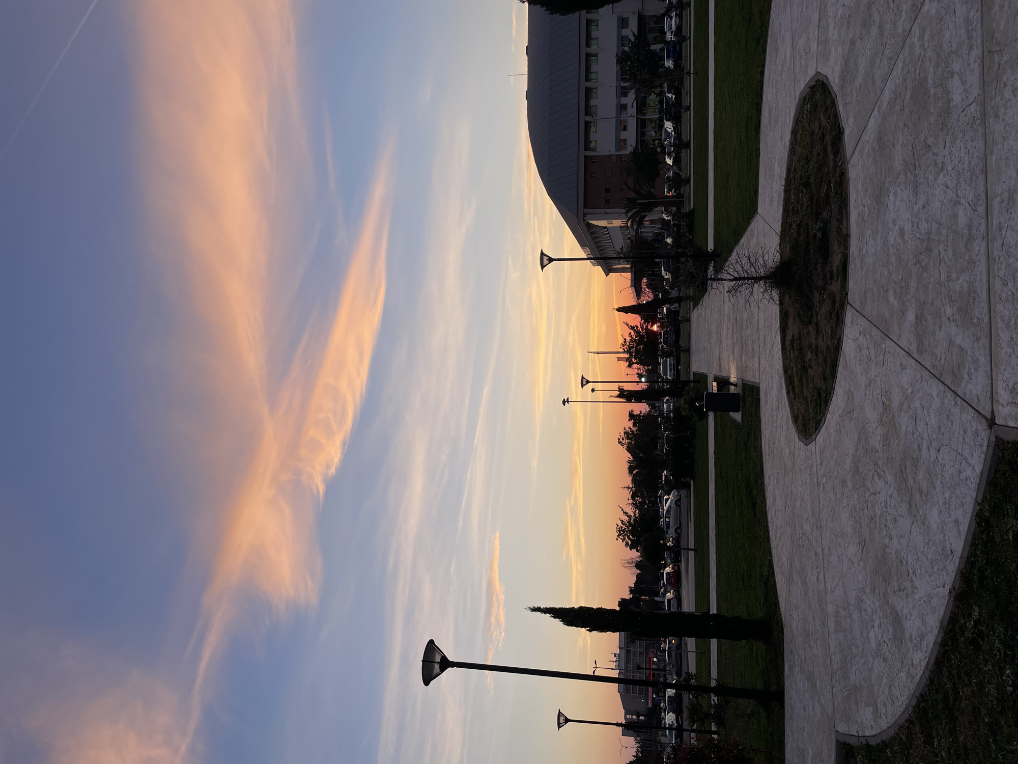 The image displays an outdoor urban environment at dusk, specifically a plaza or park area in Bar, Montenegro, located adjacent to the Sports Center "Topolica" building. The scene faces west towards the setting sun.
The sky occupies the upper two-thirds of the frame, transitioning from a pale blue-purple at the top to vibrant orange and pink hues near the horizon. Elongated, wispy clouds, illuminated in shades of orange and pink, stretch across the sky. A faint, linear contrail is visible in the upper right.
In the immediate foreground, a light-grey concrete pathway with a patterned texture curves across the bottom, encompassing a small, circular planter bed containing dark soil and a single small, bare deciduous tree. Green grass borders the pathway.
Three tall, dark streetlamps with wide, flat tops are positioned: one prominently on the mid-left, a second further back on the left, and a third on the mid-right. To the left of the pathway, a slender, dark green coniferous tree, resembling a cypress, stands upright.
The mid-right of the image is dominated by the Sports Center "Topolica," a large, modern building featuring a distinctive dark-grey, curved, domed roof. Its lower levels are lighter in color with multiple rectangular windows.
In the background, beyond a line of various trees and shrubs (including palm trees), two separate parking areas are visible, containing numerous parked vehicles of various models and colors. The vivid orange disc of the setting sun is partially obscured by trees and distant structures on the central-left horizon. Further back on the left, several multi-story urban buildings are silhouetted, one exhibiting a partial "HOTEL" sign. No individuals or specific actions are discernible within the frame.