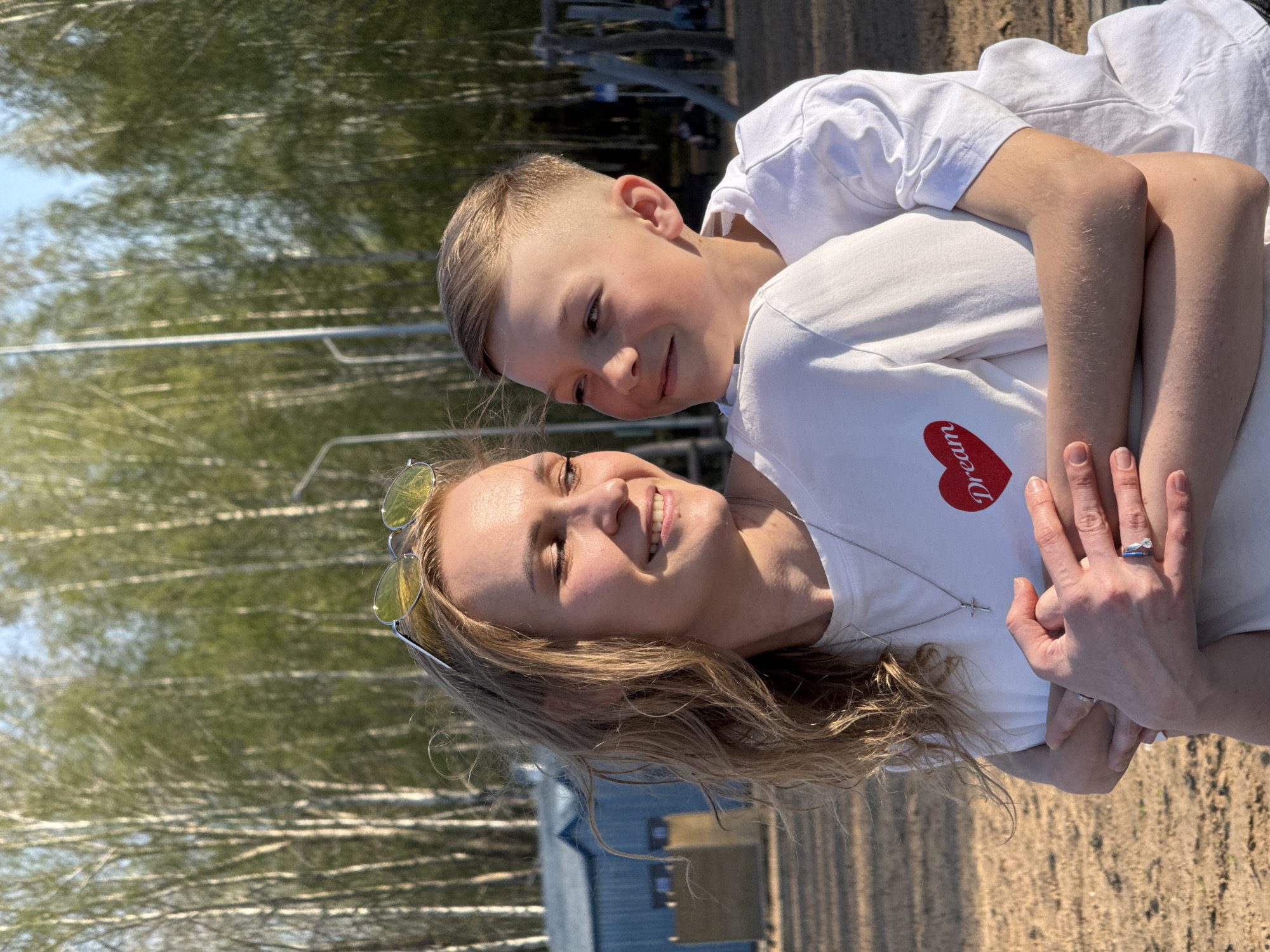 A sunlit, outdoor scene depicts a smiling woman with long blond hair embracing a young boy, likely her son. Both are wearing white t-shirts. The woman's shirt has a red heart design with the word "Dream" inside. The boy has a short haircut and is also smiling. The woman's sunglasses are perched on her head. She wears a wedding ring and a silver necklace with a cross. The backdrop consists of tall, slender trees, likely birch, and a blue building.
While the request mentioned the city of Chaikovsky, Russia, there's nothing visible in the image to definitively confirm that specific location. The landscape with the trees suggests a park or outdoor recreational area.