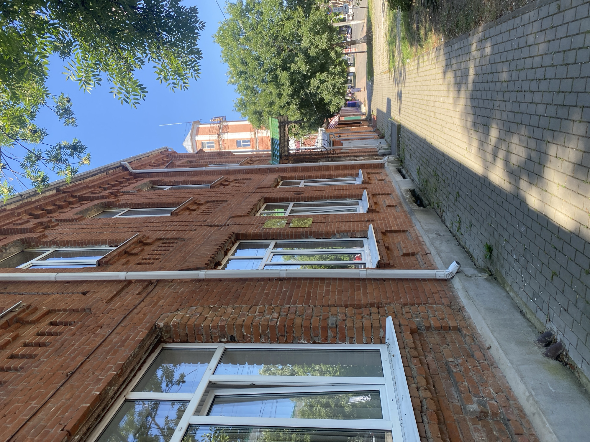 The image depicts a street scene in Maykop, Russia, on a sunny day. A red brick building with white-framed windows lines the left side of the frame. A paved sidewalk runs alongside the building, with shadows cast by trees overhead. In the background, a tall, red and white tower-like structure is visible, along with other buildings and green trees. A few people are walking on the sidewalk in the distance. The overall impression is one of a typical urban street in the summer.