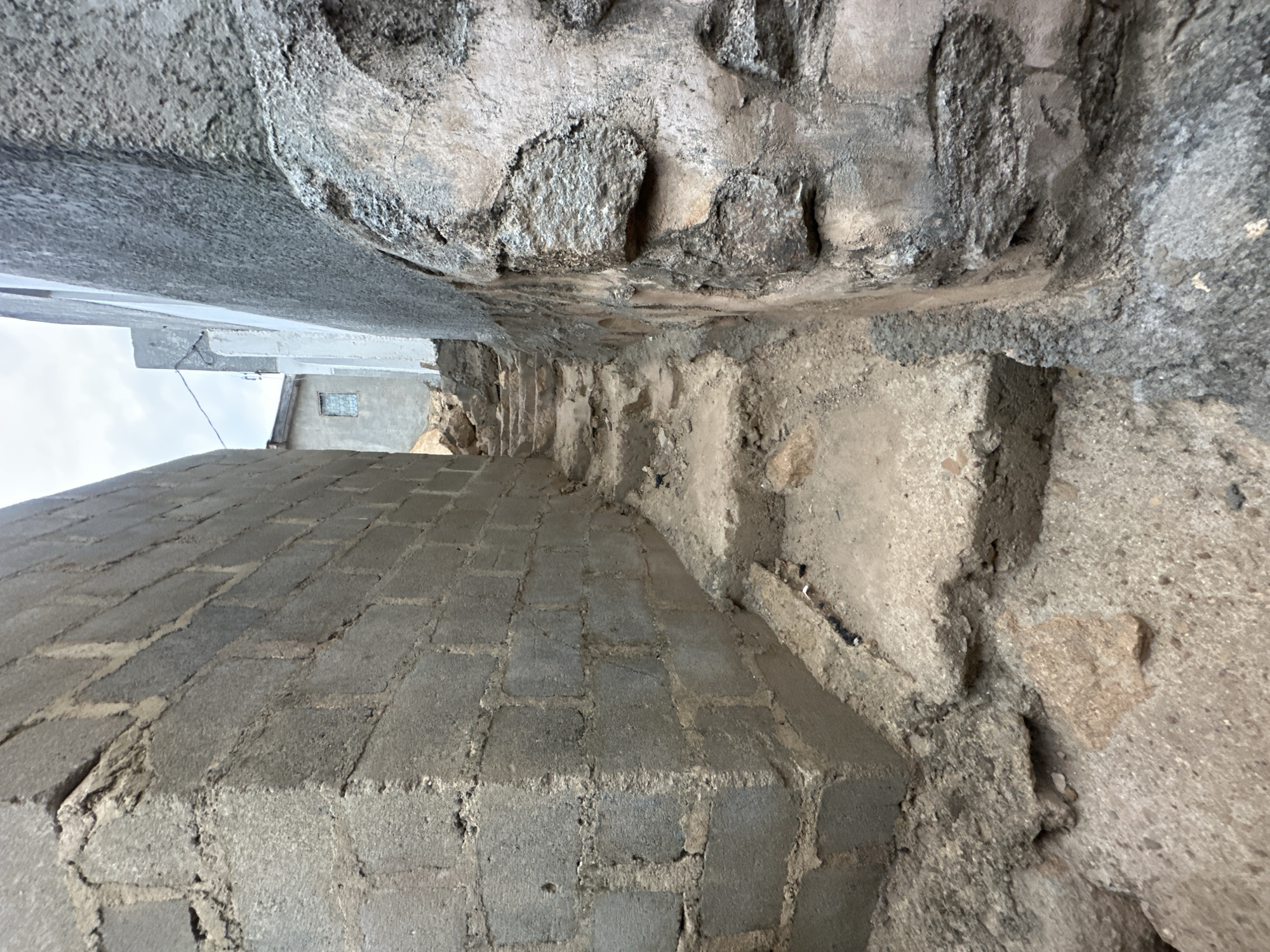 The image depicts a narrow alleyway in Jos, Nigeria. The alley is enclosed by rough stone walls on either side, topped by sections of buildings. The ground is paved with rectangular concrete blocks. There are no people visible in the image. A building with a small window is seen at the far end of the alley. The perspective is unusual, taken from ground level looking upwards.