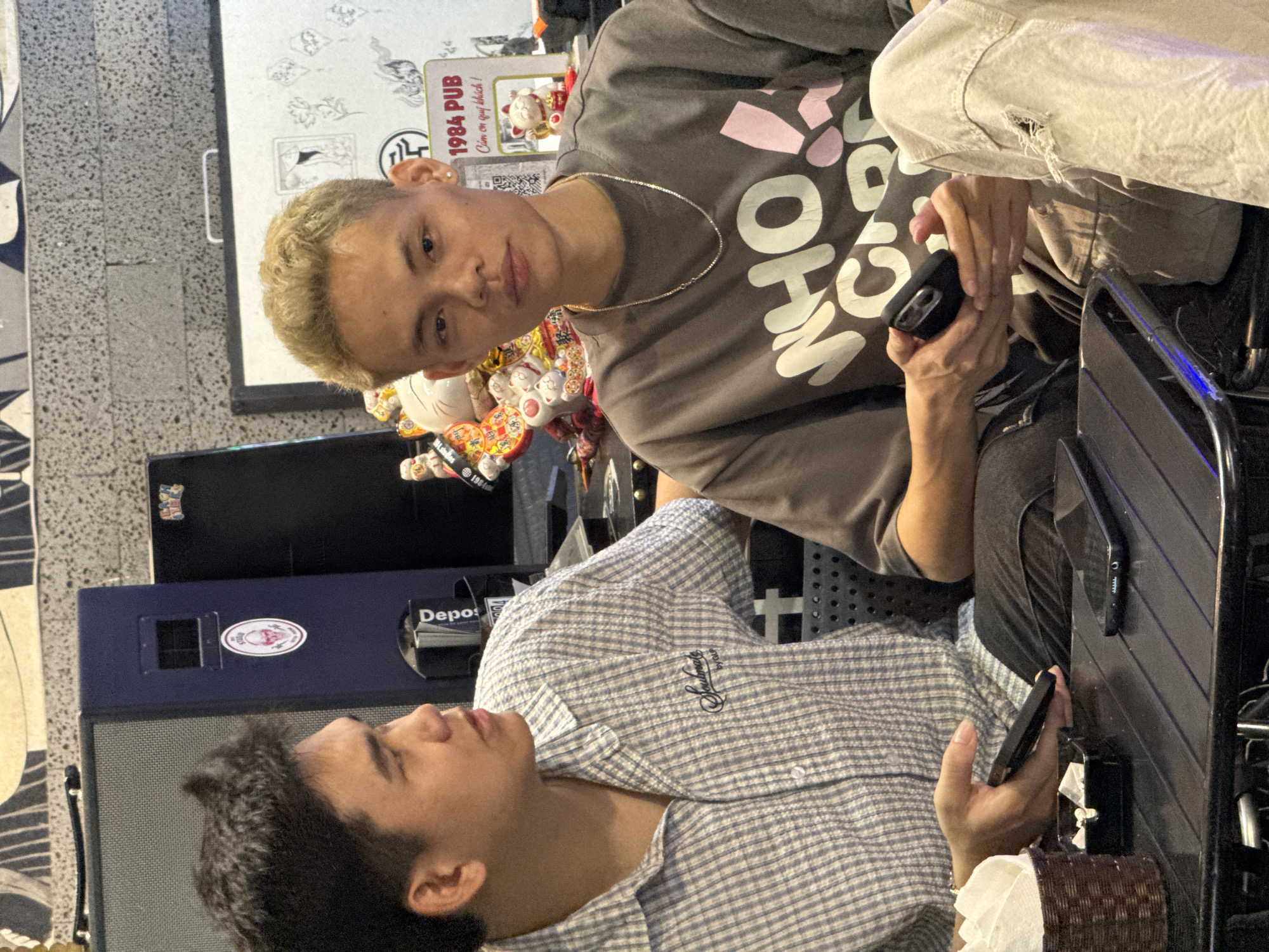 The image shows two young men sitting at a table, likely in a pub or cafe in Hanoi, Vietnam. The man on the left, with dark hair, is wearing a checkered shirt and looking to the side. He is holding a dark-colored phone in his left hand. The man on the right, with blonde hair, is looking directly at the camera. He is wearing a gray t-shirt with the text "WHO CARE" printed on it and has a silver chain around his neck. He is holding a black phone in his hands. On the table, there are two phones, a napkin holder with white napkins, and a small black ashtray. In the background, there are speakers, some decorations including maneki-neko cats, and a sign that reads "1984 PUB" and "Cảm ơn quý khách!". The overall environment appears to be casual and social.