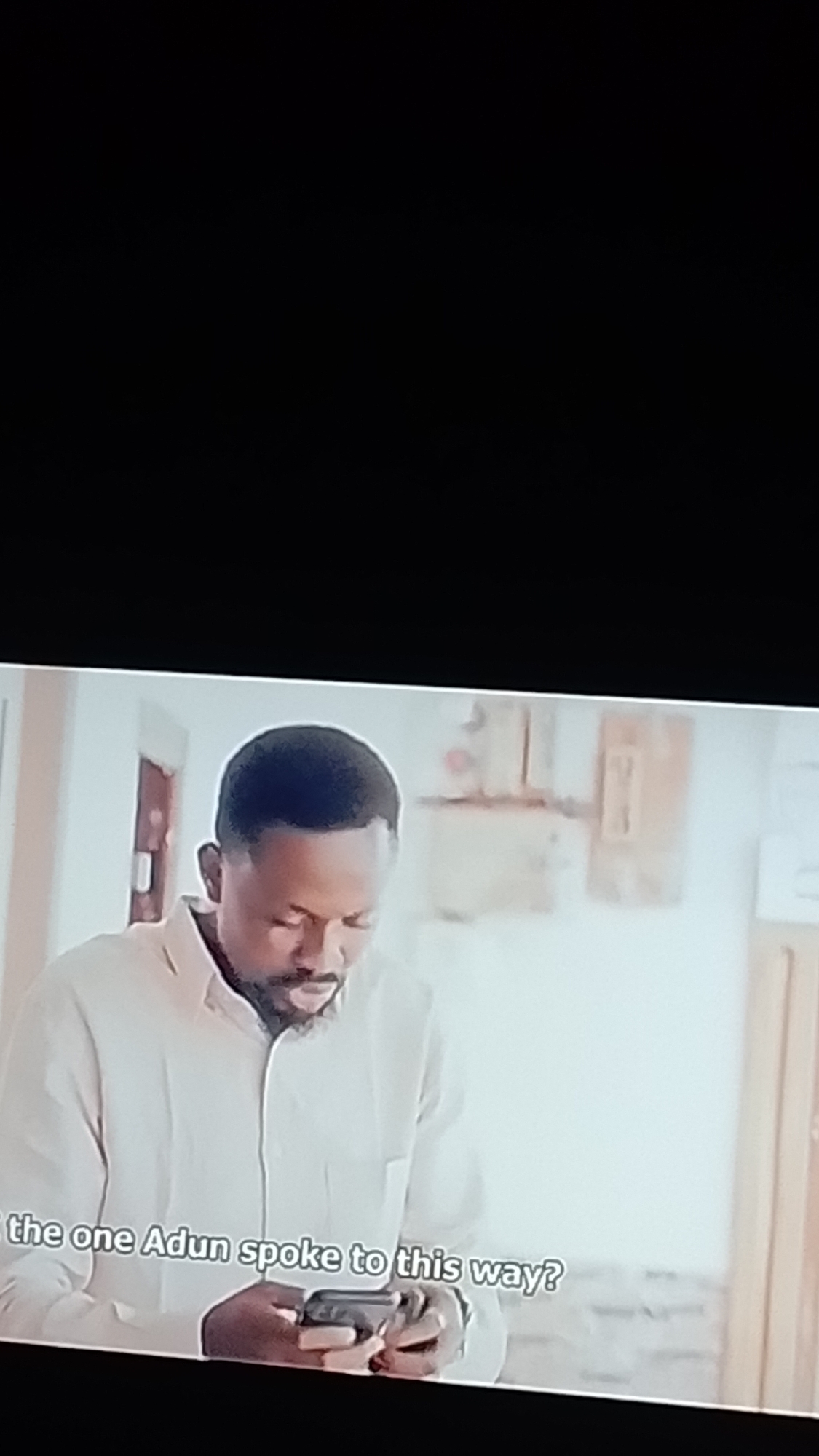 The image shows a medium shot of a Black man, likely in his late 30s or early 40s, wearing a light-colored collared shirt. He has a short beard and is looking down at a smartphone he is holding with both hands. The man is positioned in the foreground, with his upper body and the phone clearly visible. The background is blurred but appears to be an indoor setting, possibly a home or an office, with hints of a doorway and wall decor. The lighting is soft, suggesting an indoor environment. A caption at the bottom of the image reads, "the one Adun spoke to this way?". There is no visible indication of the city of Lokoja, Nigeria, in the image itself.