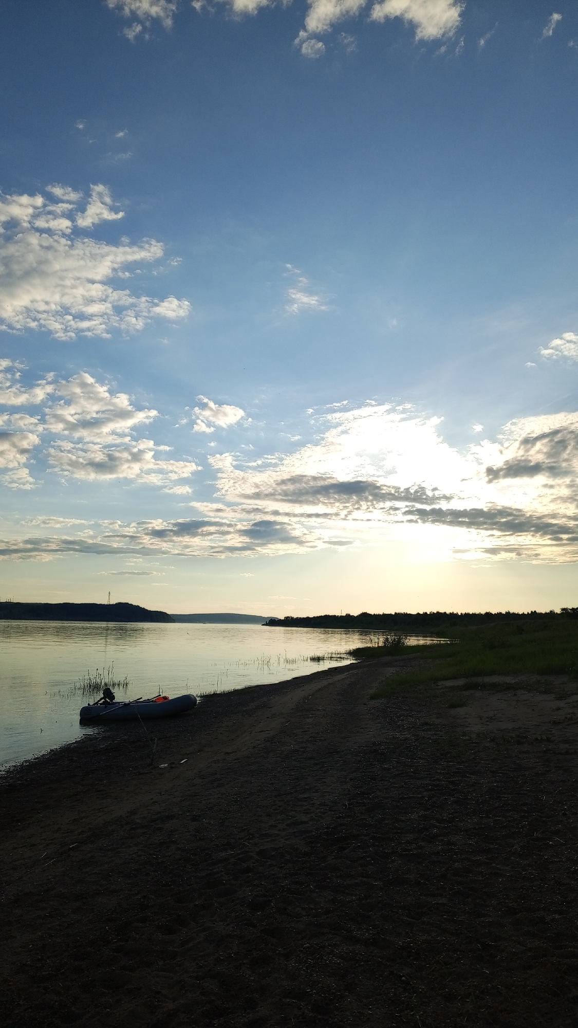 The image presents a wide outdoor vista captured during dusk or dawn in Шолья, Russia. The upper two-thirds of the frame is dominated by a clear blue sky interspersed with numerous white stratocumulus clouds, which are particularly dense on the left side and around the sun on the right. The sun, partially obscured by clouds, appears as an intense light source on the right horizon, creating a bright glare and discernible sunbeams. A calm body of water, likely a river or large lake, occupies the mid-ground, reflecting the ambient light and showing minimal surface disturbance. On the distant left bank of the water, a landmass or hill is visible, topped by a distinct, silhouetted structure, possibly a transmission tower. The immediate foreground features a dark, sandy beach curving from the bottom left to the mid-right. A grey inflatable boat with an attached outboard motor rests partially beached on the sand and partially in the shallow water on the left-central side of the foreground. An orange object is visible inside the boat. Thin reeds or tall grass protrude from the water along the near shoreline. The right side of the sandy bank transitions into patches of green grass and low vegetation, leading to a dense line of trees forming the distant right bank. No human figures are discernible in the scene.