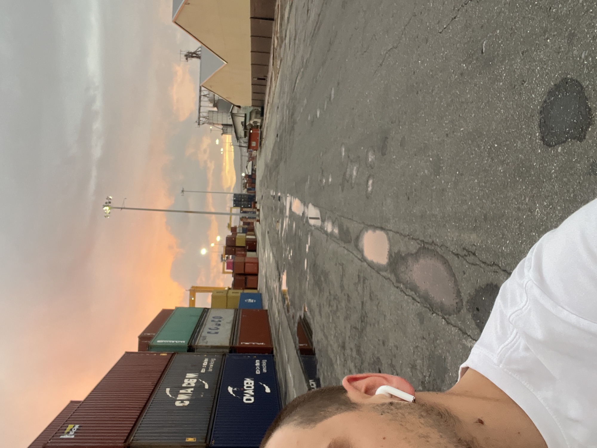 The image shows an individual wearing a white shirt and an earbud in their right ear, with stacks of shipping containers on the left. The background includes a grey concrete surface and a view of buildings and utility poles against an evening sky with orange hues. The location appears to be a shipping yard.