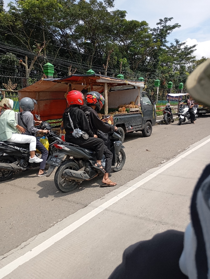 The scene shows a street in Kutawaringin, Indonesia, with a truck parked on the right side of the road, selling fruit. There are several motorcycles parked in front of the truck, with three people on the motorcycles. The person on the far left motorcycle is a woman wearing a white shirt and a green hijab. The person on the second motorcycle from the left is a man wearing a black shirt. The person on the right motorcycle is a man wearing a red helmet. There is a man wearing a white helmet on the truck, facing the back of the truck. The truck is facing forward and has a wooden structure in the back, which appears to be a stall selling fruit. There is a white line separating the right side of the road from the left side, with the truck parked on the right side of the road. The environment is clear and sunny, with a blue sky and some vegetation on both sides of the road. The image is taken from the perspective of a person sitting on another motorcycle, facing forward. The photographer's hand holding the camera can be seen in the bottom left corner.