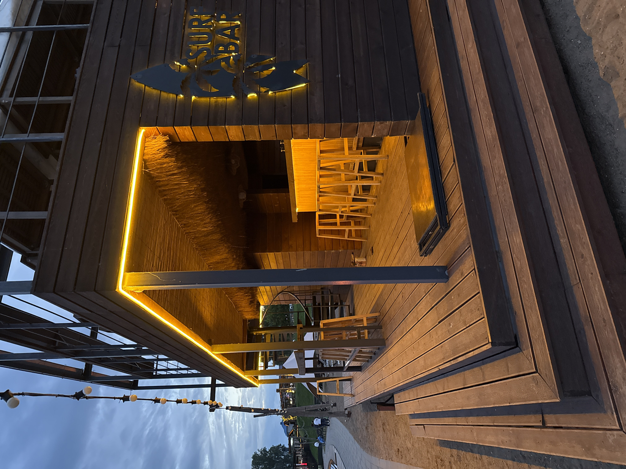 This image depicts the exterior of a wooden, multi-level establishment referred to as "SURF BAR" in Votkinsk, Russia. The structure features dark wood siding and a thatched roof over a bar area. Yellow LED lights illuminate the edges of the upper levels and the bar counter, where several wooden stools are visible. A spiral staircase leads to an upper deck with a railing. Wooden decking forms a patio area with steps leading down to a sandy or gravelly ground. In the background, other buildings, trees, and a cloudy sky are visible. Several people are faintly visible in the distance on a paved walkway. The scene appears to be set during twilight.
