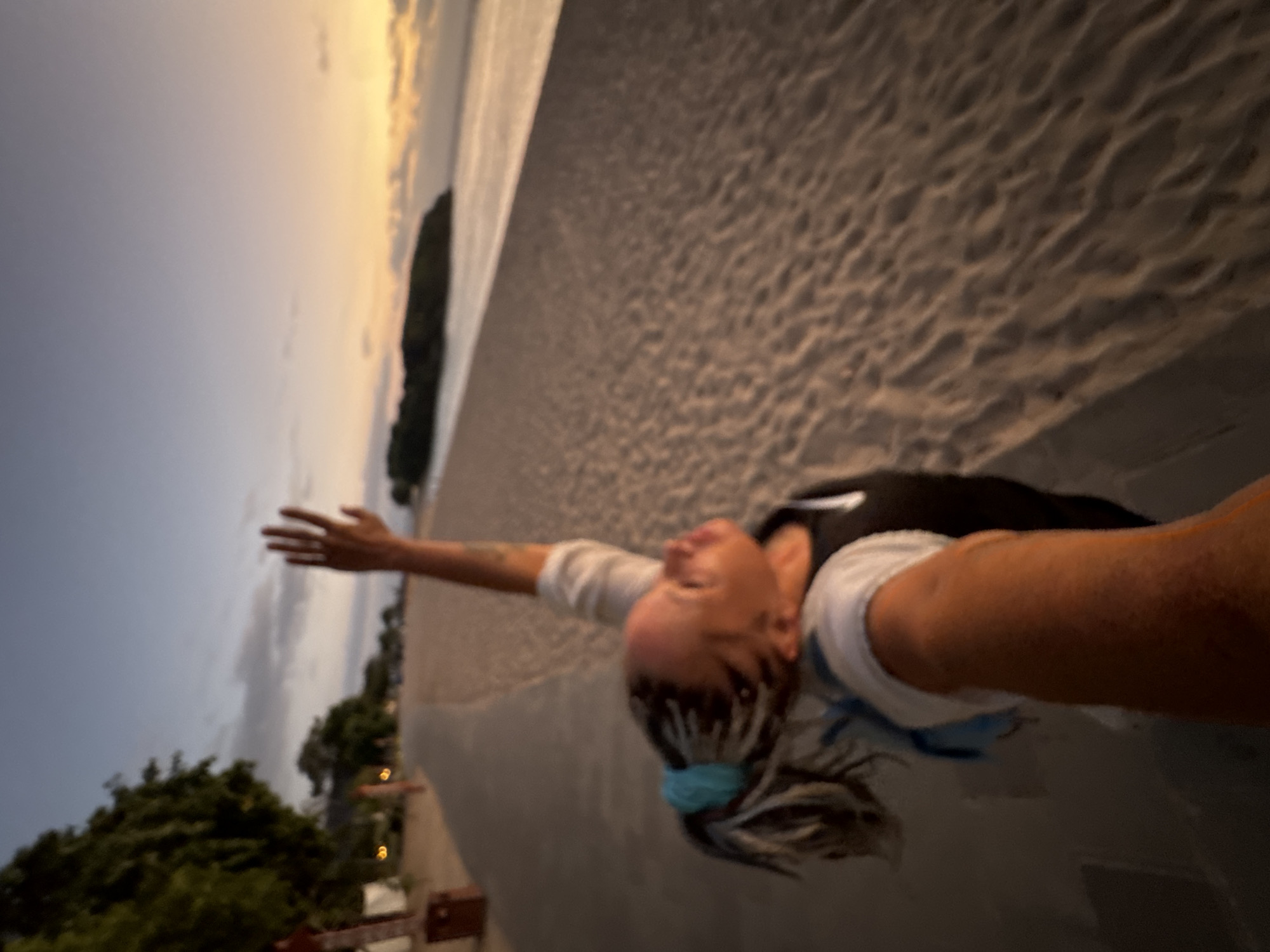 A woman is captured in a dynamic pose, leaning back with outstretched arms and head tilted upwards, eyes closed or looking towards the sky, suggesting a moment of joy or freedom. She wears a white t-shirt, a dark top layered over it, and has light-colored hair, possibly braided or dreadlocked, pulled back with a light blue hair tie.
The scene is a coastal landscape, likely a beach, illuminated by the soft light of either sunrise or sunset. The sky displays a gradient of colors, transitioning from warm, golden-orange hues near the horizon, where it meets a calm body of water, to a muted blue in the upper parts. A dark, elongated landmass or island is silhouetted against the bright horizon in the distance. The foreground features textured sand, indicating a beach, next to a paved or concrete edge where the woman is positioned. To the left, lush green trees and faint artificial lights are visible. The overall ambiance is tranquil and serene, characteristic of twilight. There is no visible text in the image. The location is Kuta Selatan, Indonesia.