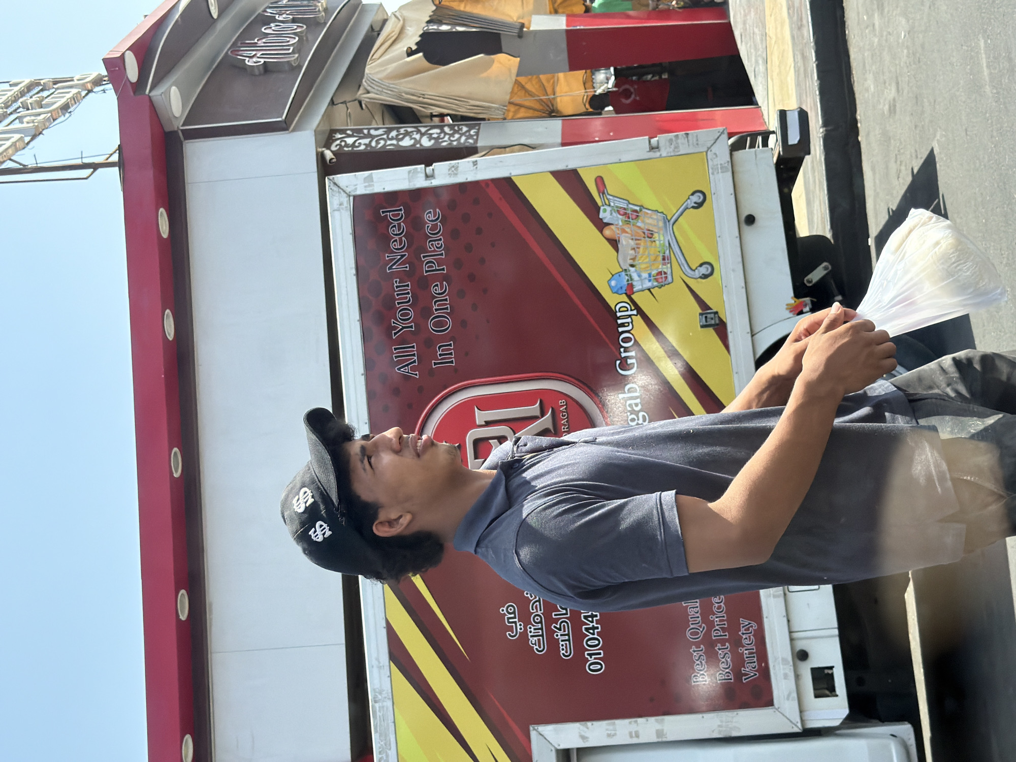 A young man wearing a dark grey polo shirt and a black baseball cap with white dollar signs is walking. He is holding a clear plastic bag filled with food in his right hand. Behind him is the side of a white truck with a red and yellow graphic design. The graphic includes the text "All Your Need In One Place" and a drawing of a shopping cart full of groceries. The truck also has Arabic text and a phone number "01044". The scene appears to be outdoors on a sunny day, possibly in a commercial area of Sharm El Sheikh, Egypt, given the presence of shops and signage in Arabic.