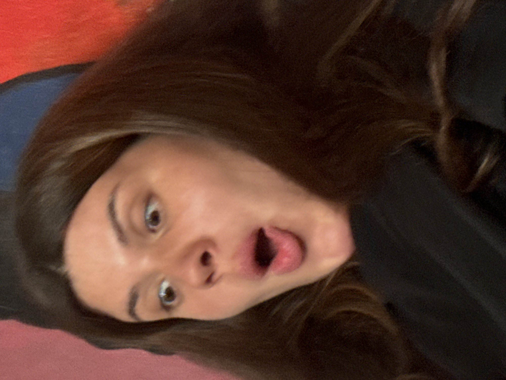 A close-up portrait depicts a young person's face, oriented sideways with the head rotated approximately 90 degrees counter-clockwise. The individual has long, dark brown hair that frames their face and extends into the background. Their eyes are wide open, conveying a sense of surprise or alarm, and their mouth is open in a large 'O' shape, with the tongue partially visible. The facial expression suggests either a strong emotional reaction or an intentional, playful pose.
The background features various colored sections. To the upper left of the head (when oriented upright), a vibrant red surface is visible, adjacent to a blue strip. To the right and below the head, there is a dark, possibly black, fabric or material, which might be part of clothing. The lighting appears to be indoor, and no specific time of day or weather conditions are discernible. There is no visible text in the image. The location context provided is Krasnoyarsk, Russia.