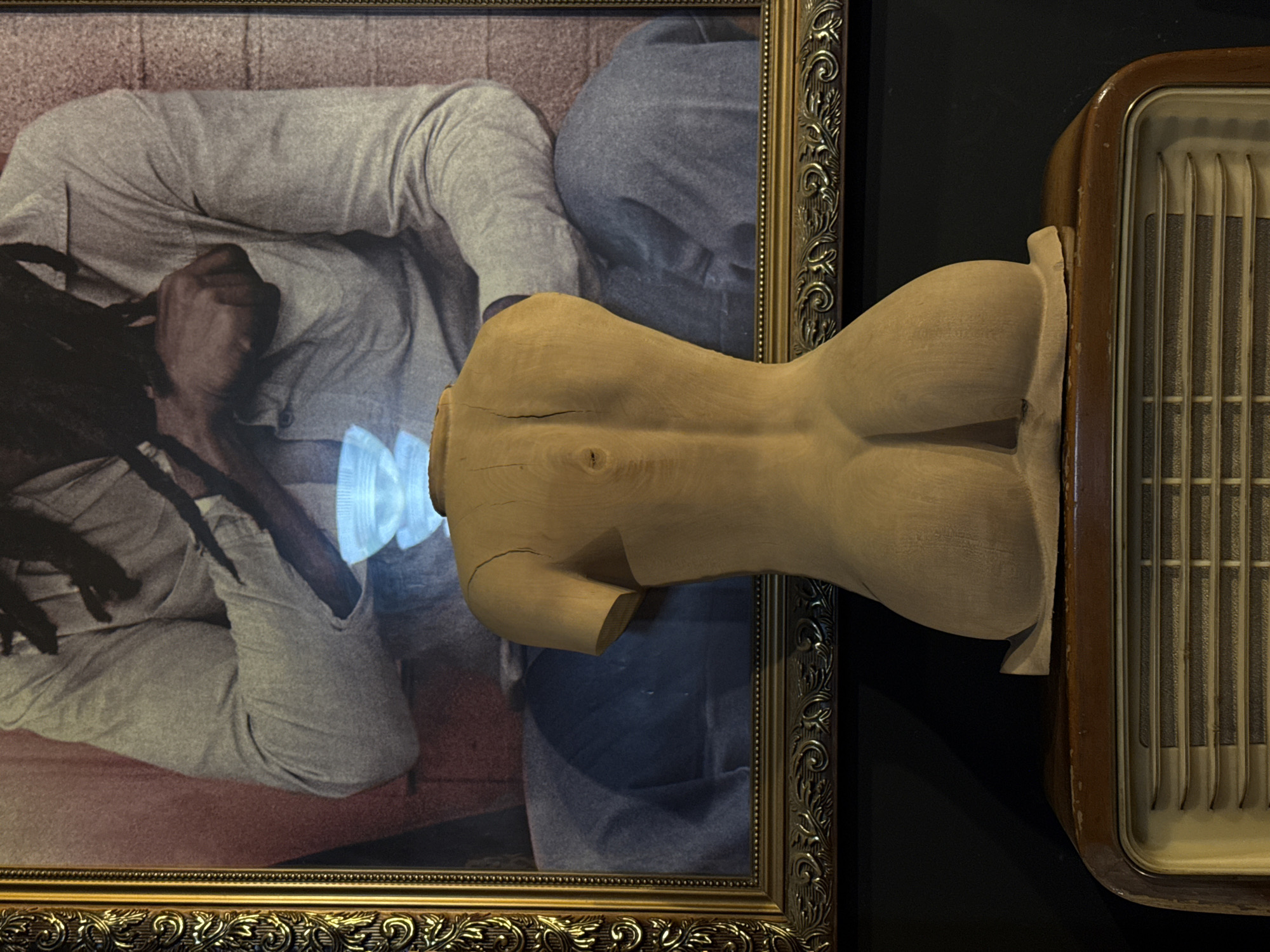 The image presents a still life composition featuring several elements. In the background, a framed picture displays a person lying down, dressed in casual attire, with dreadlocks visible. The frame is ornate and gold-colored. In the foreground, a wooden torso sculpture is positioned. To the right of the sculpture, a vintage radio with vertical grilles is placed. The photograph may have been taken indoors, but there is not enough information to specifically indicate the city of Yerevan, Armenia.