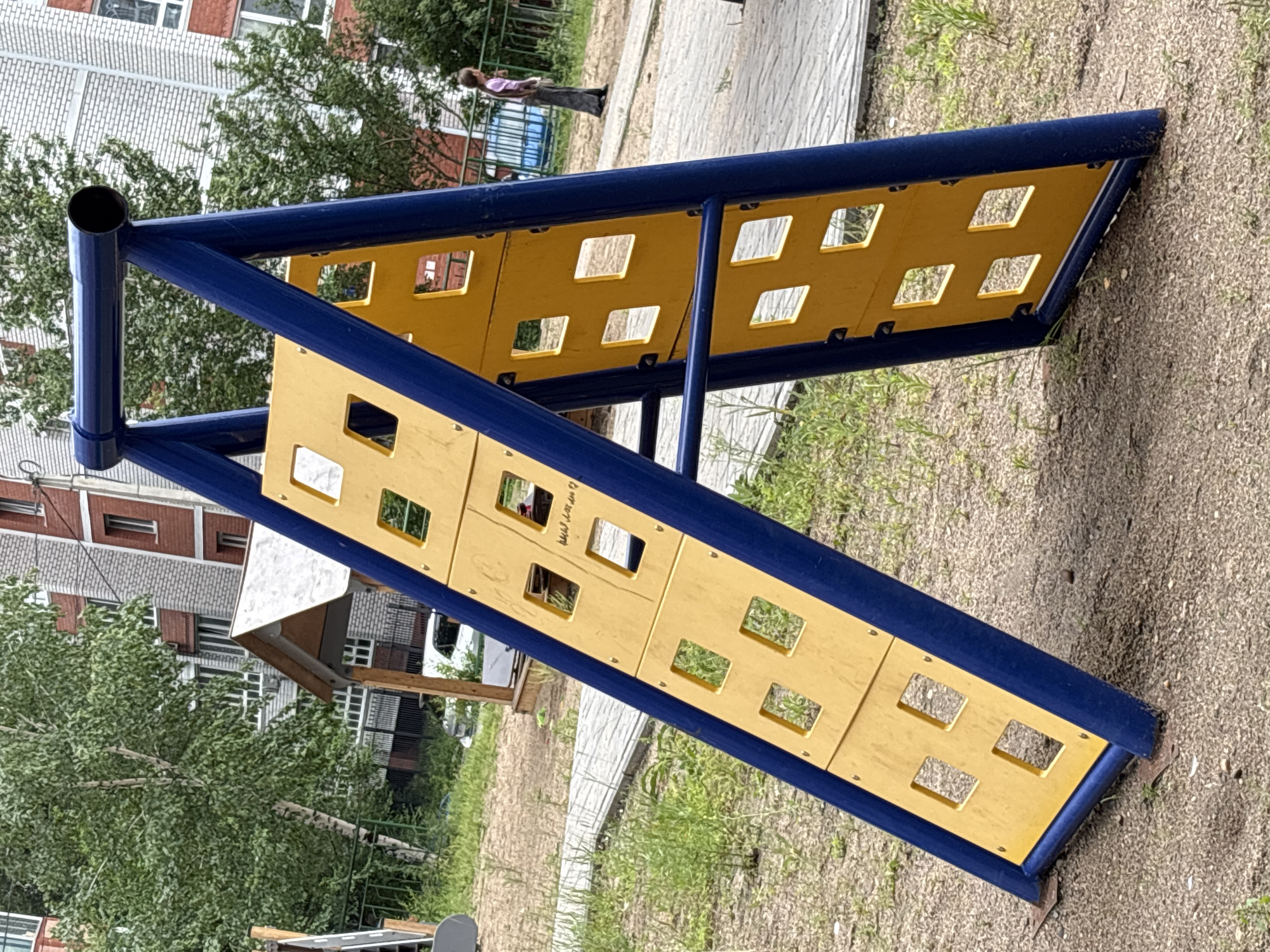 An A-frame climbing structure with blue metal supports and yellow paneling with square cutouts stands on a gravel and grass surface in a city park or playground. The structure is angled, with one side steeper than the other. In the background, there are multi-story brick apartment buildings, trees, and a paved walkway. A person in a pink shirt and dark pants stands on the walkway in the background, facing away from the camera. A small wooden structure, possibly a shed or playhouse, is visible to the left of the climbing structure, behind a metal fence. The scene appears to be in an urban residential area.