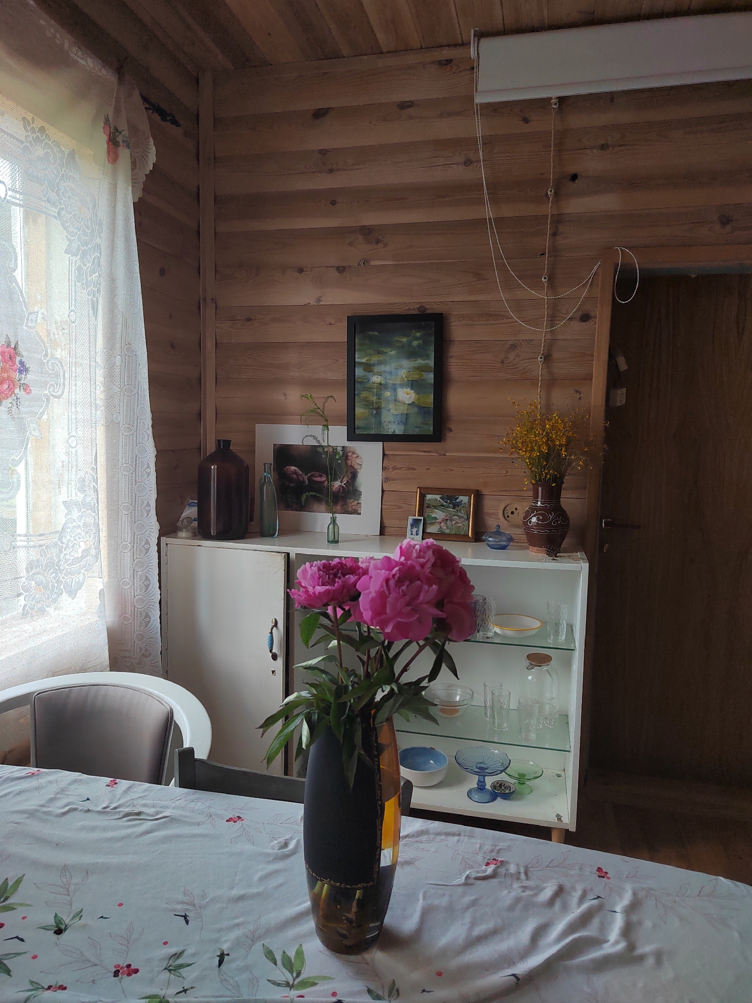 The image shows an interior scene in a room with wooden paneled walls. In the foreground, a table is covered with a white tablecloth featuring a delicate floral and bird pattern. On the table, a dark vase holds a bouquet of vibrant pink peonies. To the right, a white cabinet with glass doors displays various glassware, including bowls and pitchers. Above the cabinet, on the wooden wall, hang several framed pictures. A dark framed picture with a water scene is at the top, and a smaller framed picture with a landscape is below it. To the right of the cabinet, a brown ceramic vase holds a bouquet of yellow flowers. A closed wooden door is visible on the far right. To the left, a sheer white lace curtain is draped over a window, with floral motifs visible on it. A portion of a white upholstered chair is also visible on the left. The overall impression is of a cozy, domestic setting. The image is likely taken in a residential interior within the Бярэзінскі сельскі Савет region of Belarus, suggested by the context of the provided location. There are no people visible in the image.