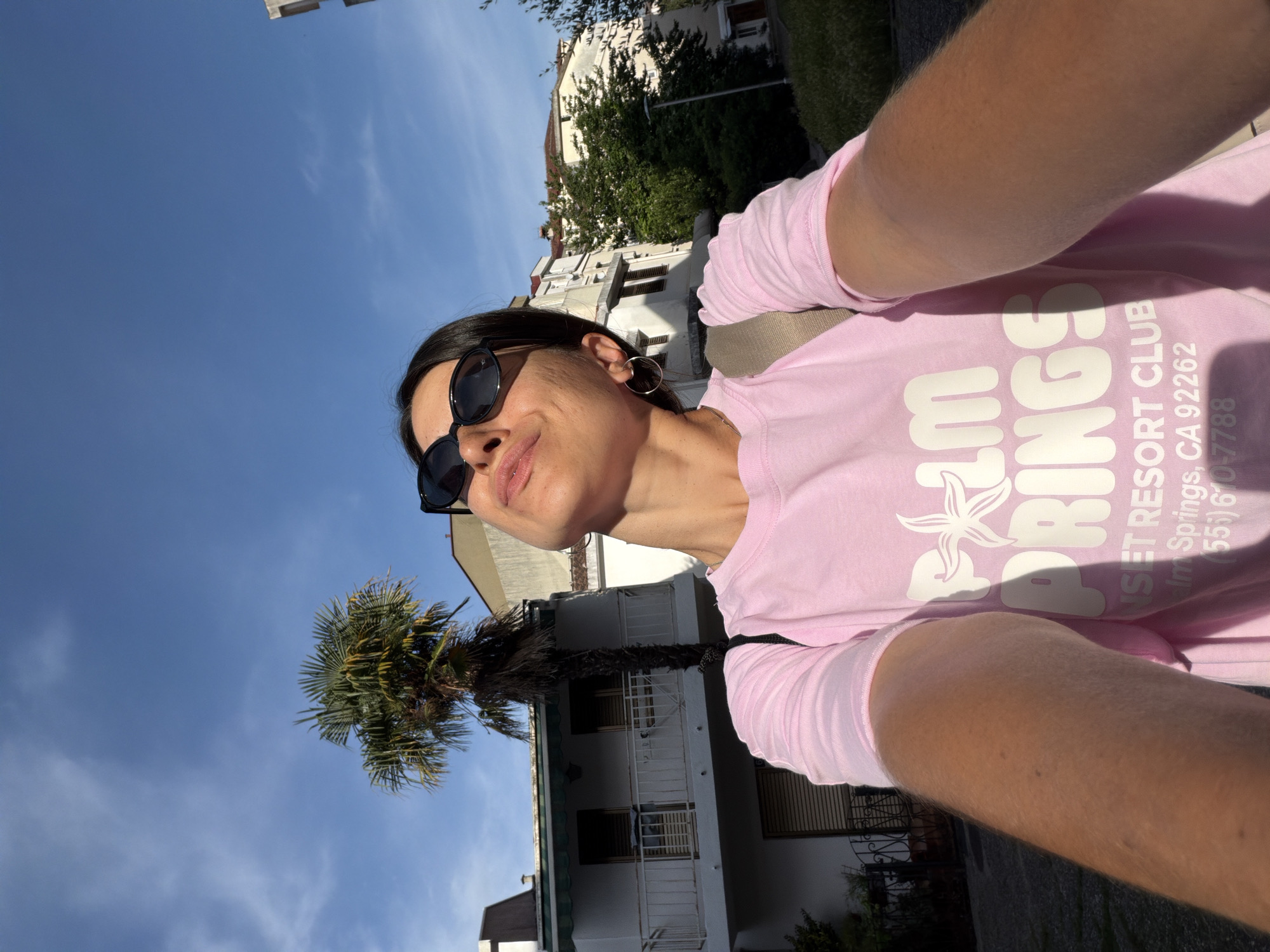 A woman wearing sunglasses and a pink long-sleeved shirt with "Palm Springs Resort Club" printed on it is taking a selfie. The background shows a clear blue sky, buildings, and a palm tree. Based on the architectural style and the presence of palm trees, the location is likely in Budva, Montenegro. The buildings appear to be residential.