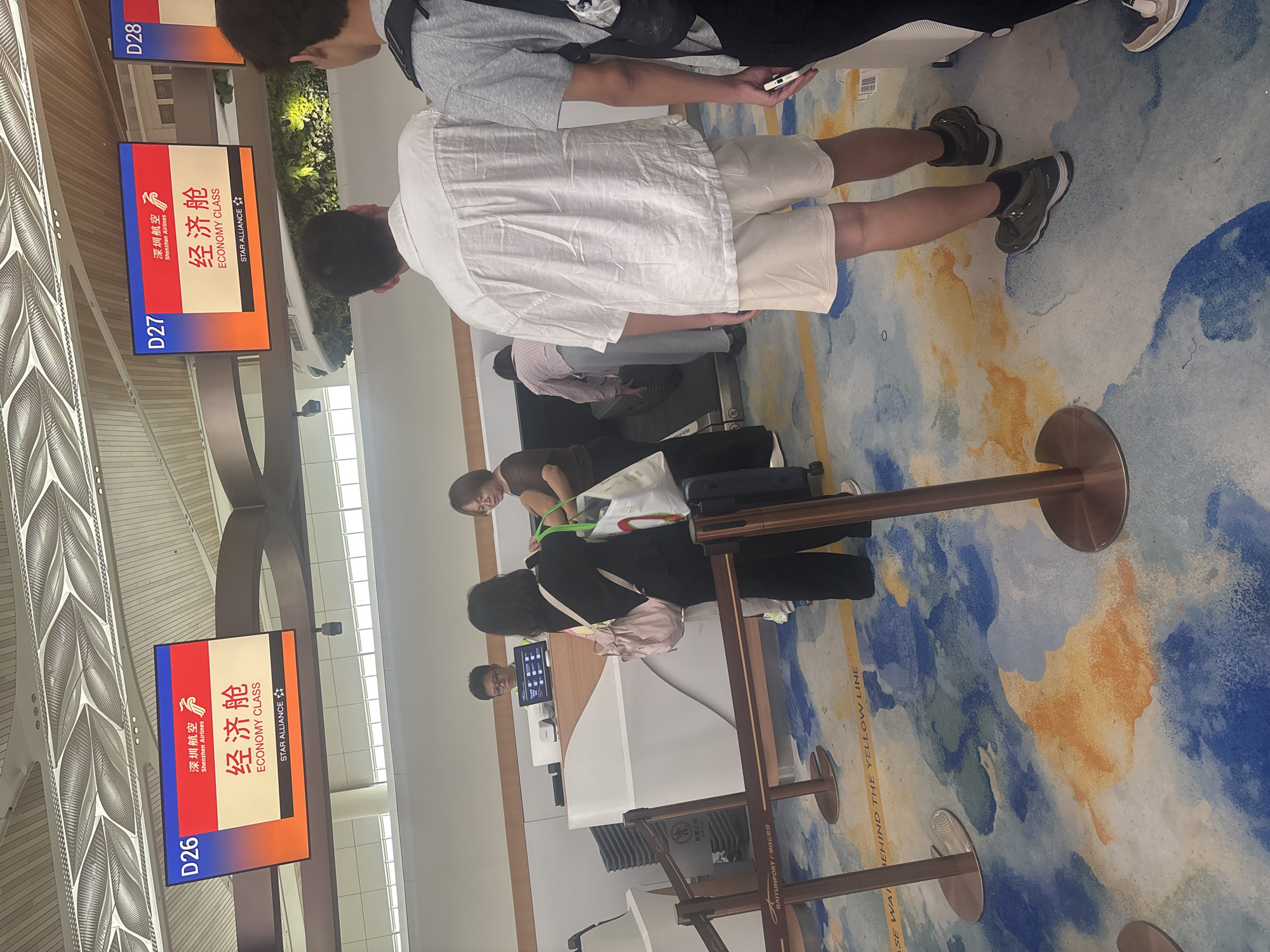 At an airport check-in counter in Baiyun District, China, passengers wait in line. Two overhead digital signs display flight information, one labeled "D26" and the other "D27," both indicating "ECONOMY CLASS" for Shenzhen Airlines. A person in a black shirt and pants is interacting with a check-in agent, who is behind a white counter. Another person in a pink and white checkered shirt is also at the counter, handling luggage. In the foreground, two individuals are standing in line, one wearing a white linen shirt and beige shorts, and another wearing a grey t-shirt and black backpack. The floor is carpeted with a colorful, abstract design featuring blues, oranges, and yellows. Stanchions with ropes guide the queue. A sign on the floor reads "PLEASE WAIT BEHIND THE YELLOW LINE." The environment appears to be an indoor terminal of an airport.