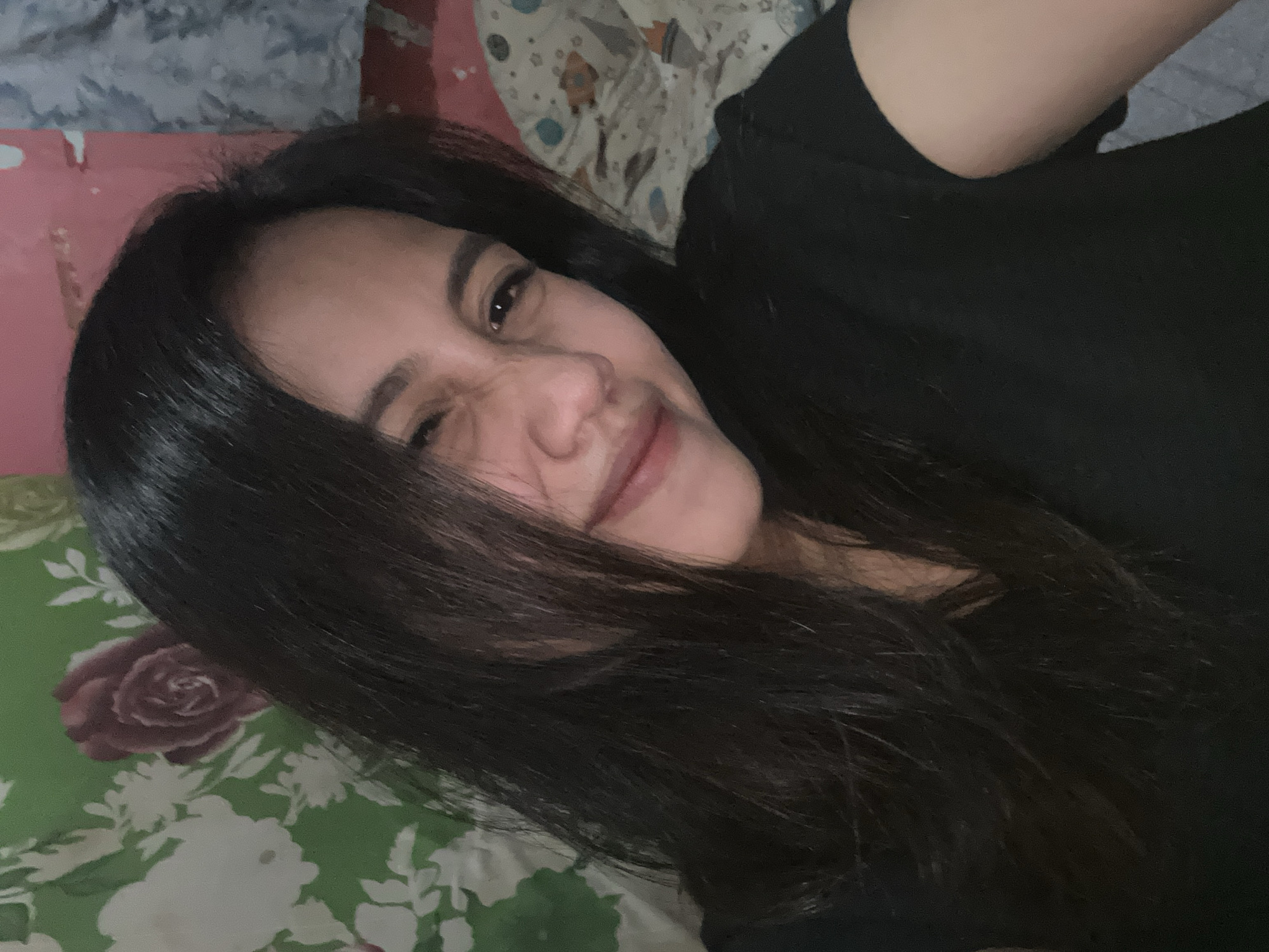 The image shows a close-up of a young woman with long, dark hair, wearing a black t-shirt. She is looking towards the camera with a playful expression, her eyes slightly squinted and her mouth partially open in a smile. Her hair partially obscures her face. The background appears to be a bedroom, with a green floral-patterned curtain or wallpaper on the left and what looks like bedding with a pattern of planets and rockets on the right. The lighting suggests an indoor setting, possibly at night or in a dimly lit room. The overall impression is of a casual, informal selfie. The image is a photograph taken in Depok, Indonesia.