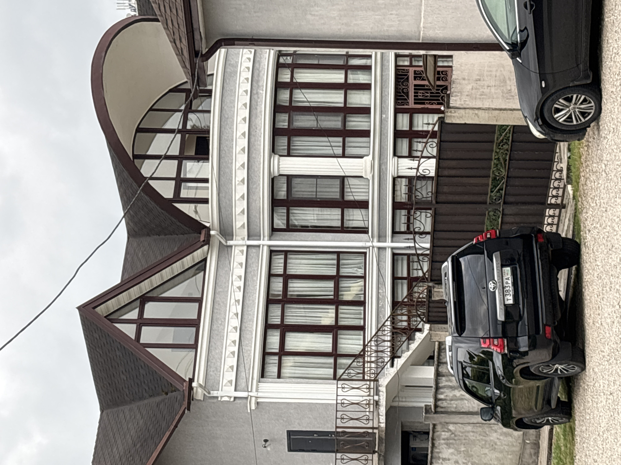 This is a vertical, slightly blurry photograph of a large, modern house with distinctive architectural features. The house is multi-story, with a light gray facade and dark brown trim around the numerous windows. The windows themselves are large and divided into many panes, with white curtains visible inside. One prominent feature is a large, curved section of the house with a rounded roofline and a large, segmented window. The roof is covered in dark shingles, and there are angular, triangular window formations on one side of the house.
In front of the house, two black SUVs are parked. The rear of one SUV, a Toyota Land Cruiser, is clearly visible, showing a license plate with the characters "T 383 PA". The other SUV is partially obscured. A dark metal fence with intricate scrollwork runs along the side of the property, and a set of outdoor stairs with a matching railing leads up to one of the entrances. The ground is covered in gravel and some patches of green grass.
The sky appears overcast, suggesting a cloudy day. There are no people visible in the image, and the scene appears to be a residential setting. The overall impression is of a somewhat imposing, contemporary home.