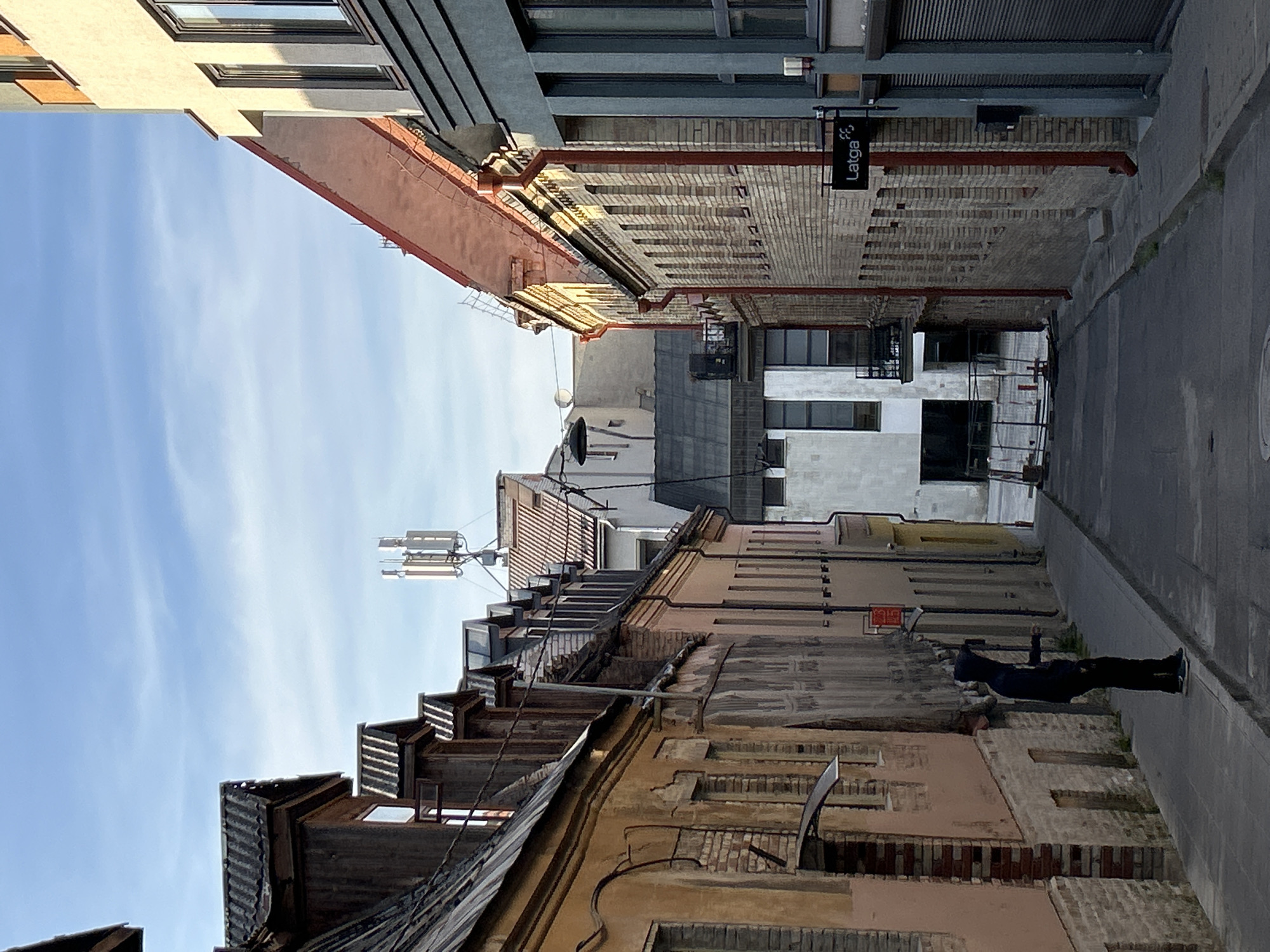 The image shows a narrow street in Vilnius, Lithuania, lined with buildings of varying architectural styles. The street slopes downwards away from the viewer. On the left, there is an older building with a beige facade, weathered brickwork, and a mansard roof. Further down the left side, there are more buildings with darker roofs and some signs of wear. On the right side of the street, a modern building with a grey facade is visible, with a sign for "Latga" attached to it. Beyond this, a building constructed of light-colored bricks rises up, with red downspouts and numerous windows. In the distance, more buildings and a telecommunications tower can be seen against a partly cloudy sky. In the foreground on the left sidewalk, a person wearing a dark hooded sweatshirt and dark pants stands facing away from the camera, with their left arm extended. The street appears to be paved with asphalt, and there are some weeds growing along the edge of the sidewalk. Construction fencing is visible further down the street in front of one of the buildings. The overall scene suggests an urban environment in a European city, with a mix of old and new structures.
