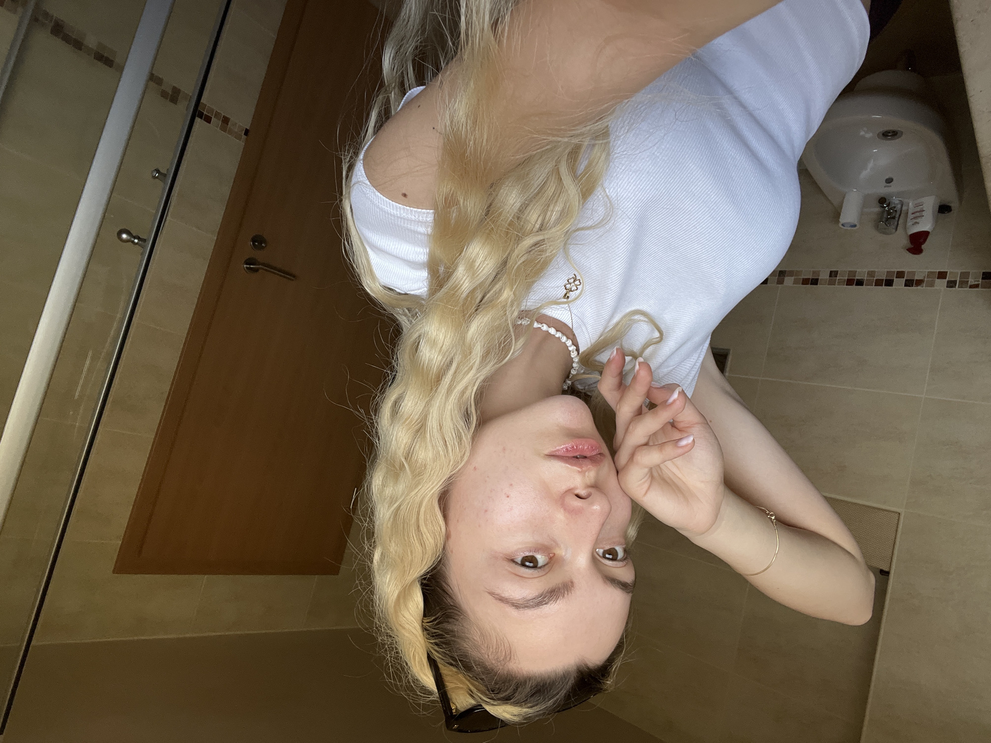 A young woman is captured in a mirror selfie within what appears to be a bathroom. She has long, wavy blonde hair with darker roots and light-colored eyes, looking directly into the camera with a neutral expression. Her face shows some minor skin blemishes. She is wearing a simple white ribbed tank top, a pearl necklace, and a thin gold bracelet on her left wrist. Dark sunglasses are pushed back into her hair. Her left hand is raised near her face, revealing light-colored nail polish.
The background elements confirm a bathroom setting. To her left, part of a shower enclosure with a glass door and chrome handles is visible, next to a wooden door with a handle. The walls and floor are covered in light-colored ceramic tiles, interspersed with a decorative horizontal band of smaller, darker tiles. To her right, a white ceramic bidet or small sink with a faucet is partially visible, on which a small red and white tube (possibly toothpaste or lotion) rests. The overall lighting is bright, suggesting daytime. No discernible text is present in the image.