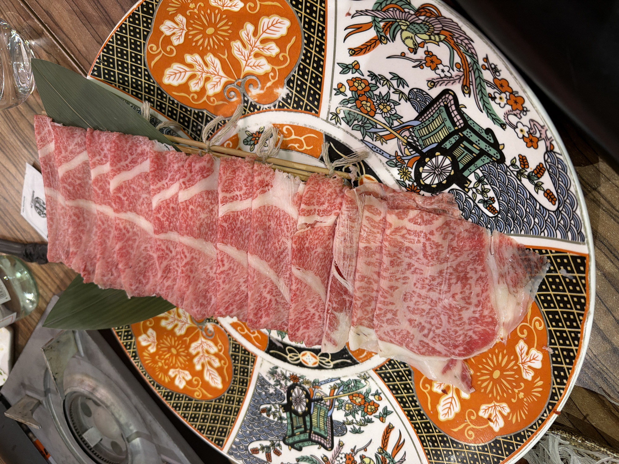 The image shows a close-up, overhead view of thinly sliced raw beef arranged on a decorative plate, with a portable gas stove and a glass of water in the background. The beef is marbled with fat and appears to be a cut for shabu-shabu or hot pot. The plate has an intricate, traditional Japanese-inspired design with floral patterns, geometric borders, and imagery of what appear to be traditional Japanese chariots or carts, rendered in vibrant colors of orange, black, white, green, and red. A single green leaf is placed beneath the beef slices. The table surface is made of wood. There is no indication of the city of Dubai, United Arab Emirates.