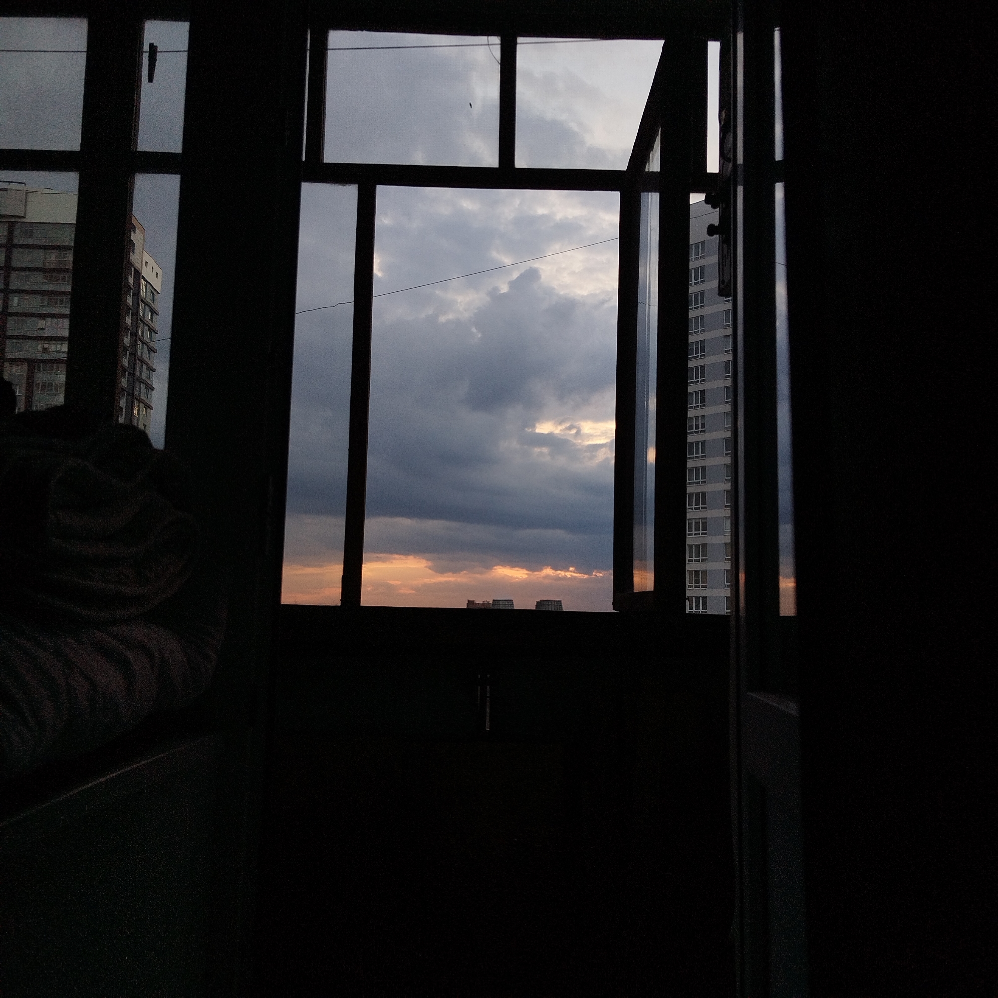 The image presents a view through a window with a grid frame, likely in a multi-story building in Yekaterinburg, Russia. Outside the window, the sky is visible with dark, ominous clouds and a strip of orange light near the horizon, indicative of either sunrise or sunset. In the periphery, adjacent to the windows, are buildings. The image is mainly dark, and what is inside the room where the image was taken is difficult to identify because of the dark conditions.