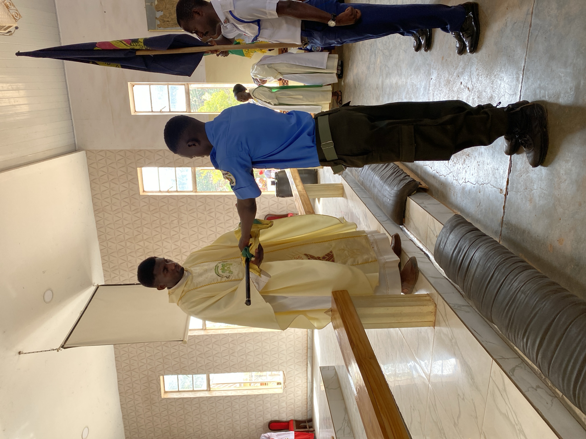 In an interior setting, presumably a church in Kuru Dungus, Nigeria, a man in a blue uniform and olive-green trousers is interacting with a clergyman. The clergyman is clad in a pale yellow robe with intricate designs and stands near a wooden structure. The uniformed man appears to be presenting something to the clergyman. In the background, there are other individuals, some in white robes and one holding a flag. The room is brightly lit with natural light streaming through windows, and the floor is tiled. The walls feature a repeating pattern.