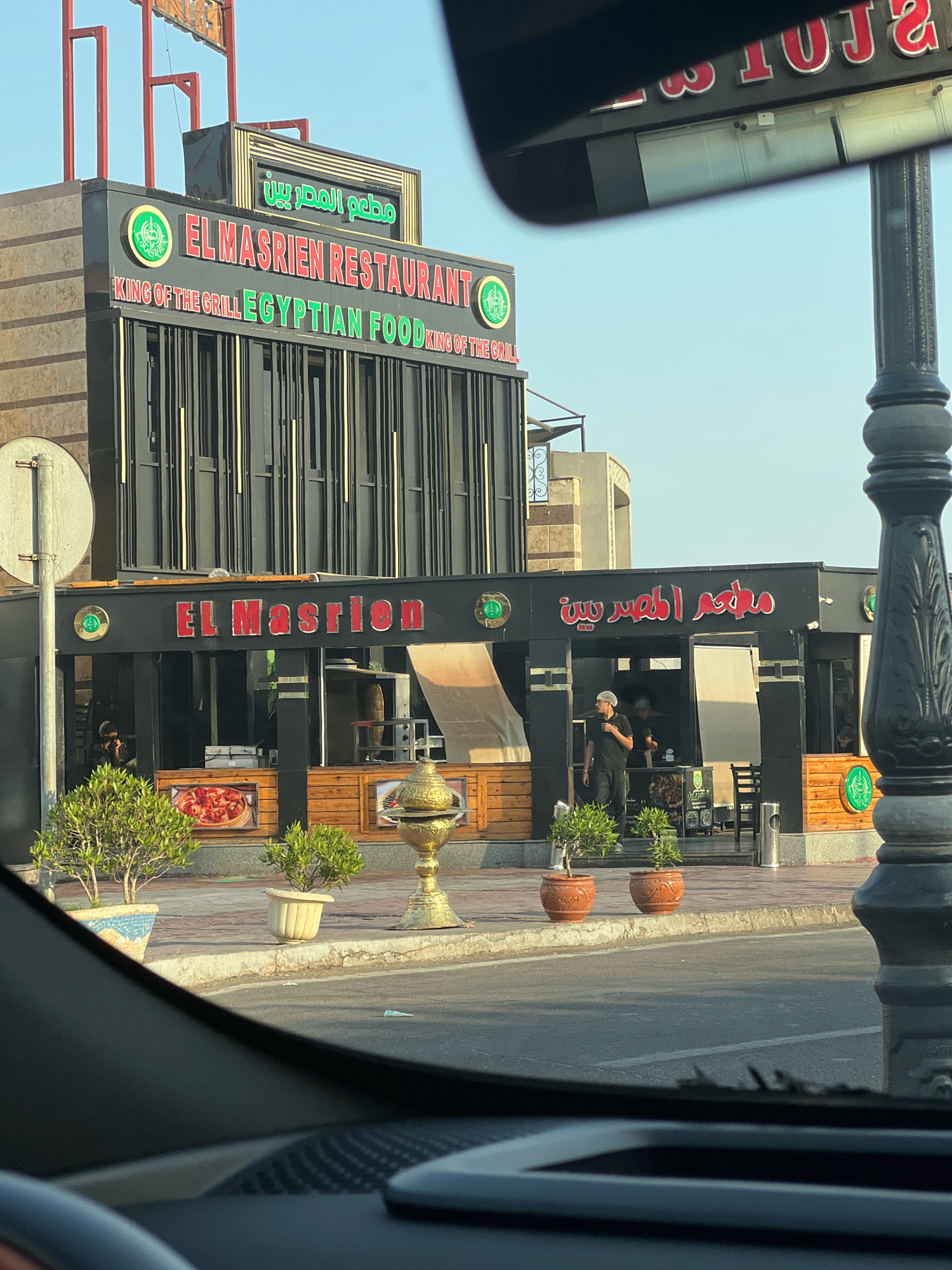 The image shows the facade of a restaurant named "El Masrien Restaurant" in Sharm El Sheikh, Egypt. The restaurant features a large sign with the name in both English and Arabic, along with the slogan "King of the Grill Egyptian Food". The building is modern with a dark exterior and wooden accents. There are windows displaying food items, and several potted plants are placed outside. Two employees are visible inside the restaurant, one in a black uniform and the other in a white cap. A lamppost stands on the right side of the image, and a street is in the foreground. The sky is clear and blue.