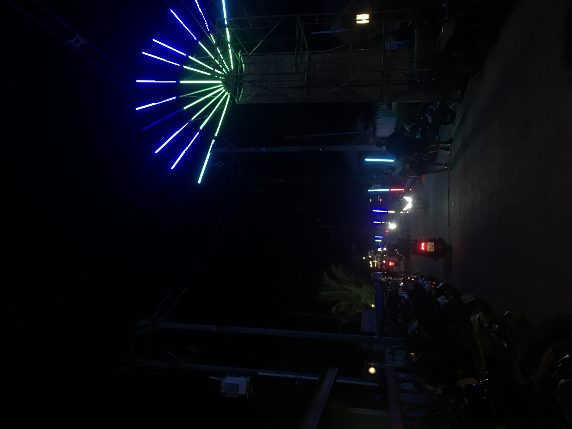 At night in Baan Mae Nam, Thailand, a street scene is illuminated by a large, decorative light installation resembling a radiant fan, with alternating blue and green light bars. The installation is mounted above the road. The street is lined with parked motorcycles, and some people are visible standing or walking along the road. The scene features the night-time environment.