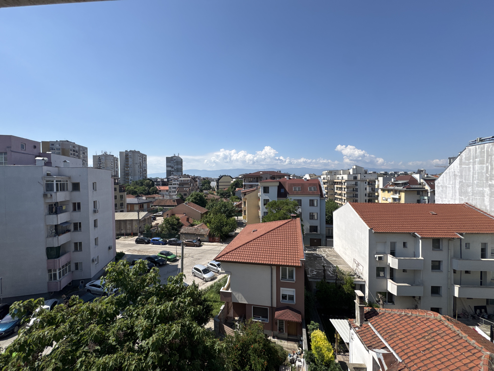 This is a daytime, eye-level shot of a residential area in Plovdiv, Bulgaria, showcasing a mix of apartment buildings and houses under a clear blue sky with scattered white clouds. The foreground is dominated by lush green trees and foliage, obscuring part of the lower section of a tall, white apartment building on the left. This building features multiple balconies and windows. Adjacent to it, a street is visible with several parked cars, including a blue sedan, a black sedan, a green hatchback, a white hatchback, and a silver sedan.
Further into the scene, a two-story house with a red tiled roof and beige and pink exterior is prominent. Several other buildings of varying heights and architectural styles, many with red tiled roofs, are scattered throughout the cityscape. In the background, taller apartment buildings and the faint outline of distant hills can be seen. The overall impression is of a typical urban residential neighborhood on a sunny day.