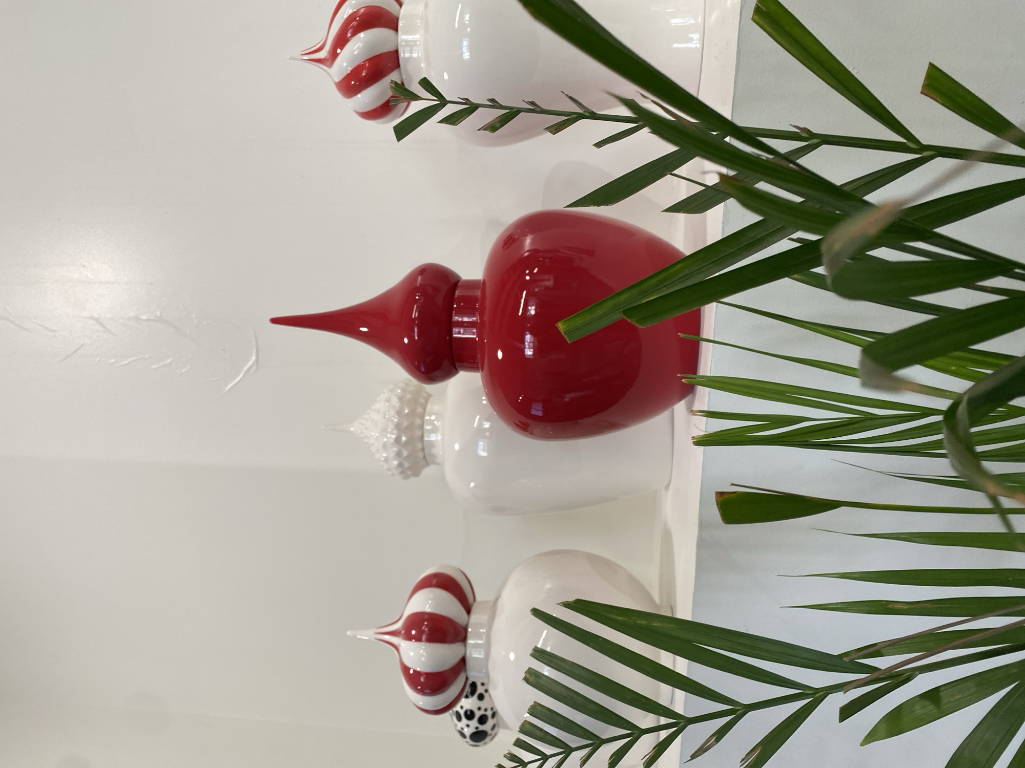 The image shows a close-up of a shelf with four decorative urns, partially obscured by green leafy foliage. The urns are all white with decorative tops. From top to bottom: the first urn has a red and white striped top; the second is entirely red; the third has a textured white top; and the fourth has a red and white striped top with a black and white polka dot base. The background is a white wall. The location and presence of people are not discernible from this image. The city of Moscow, Russia, is not identifiable in this image.