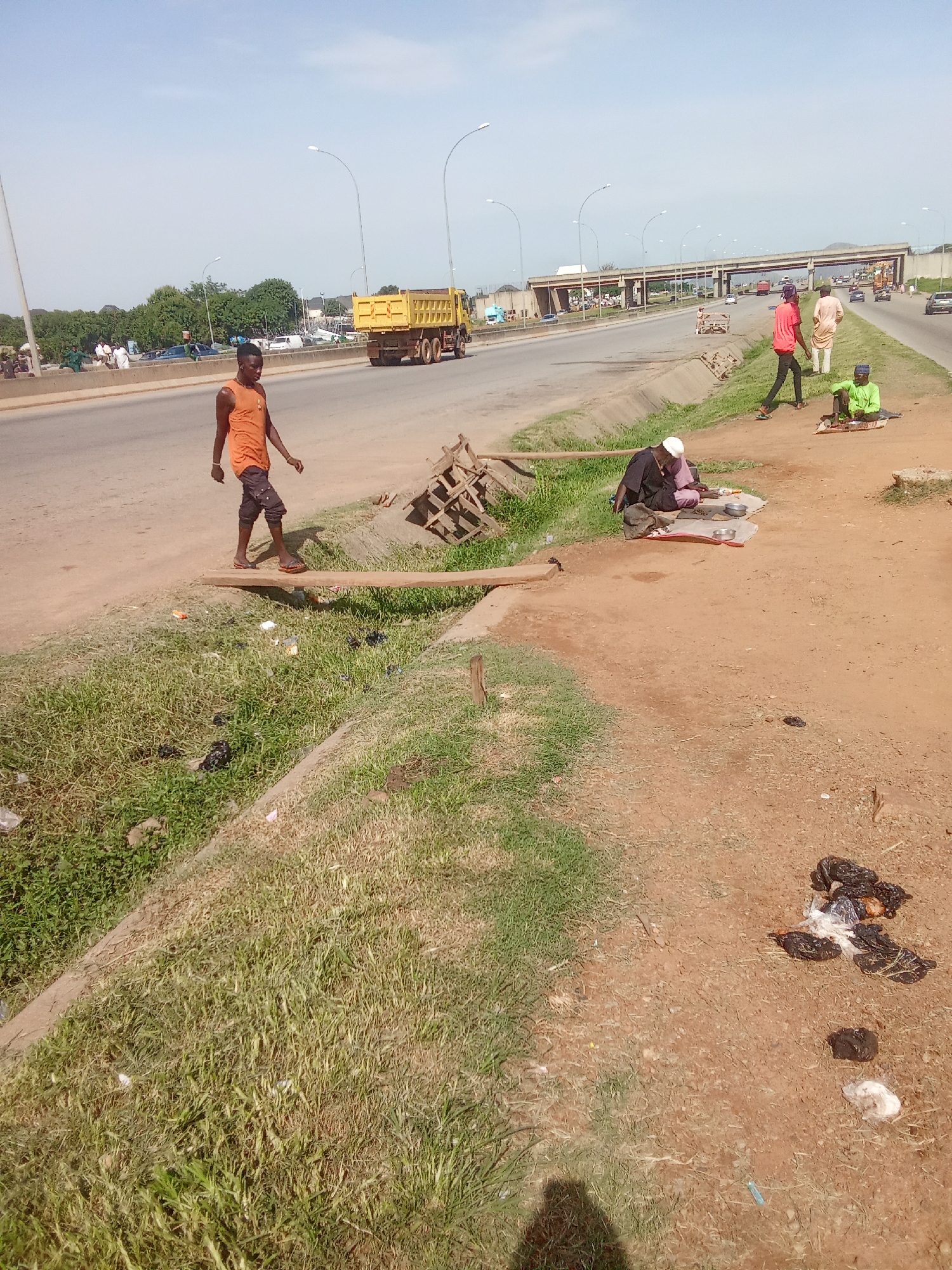 An outdoor scene depicts a roadside with several individuals and a yellow dump truck. A young man in an orange tank top and dark shorts walks across a wooden plank over a grassy ditch filled with litter. In the middle ground, a man wearing a white cap and dark clothing sits on a mat with two metal bowls and some food items. To his right, another person in a green shirt sits on a mat. Further in the background, a person in a red shirt and light-colored pants stands near a road with passing vehicles. A large yellow dump truck is visible on the road. The environment is dusty with patches of grass and debris. The sky is clear and blue.