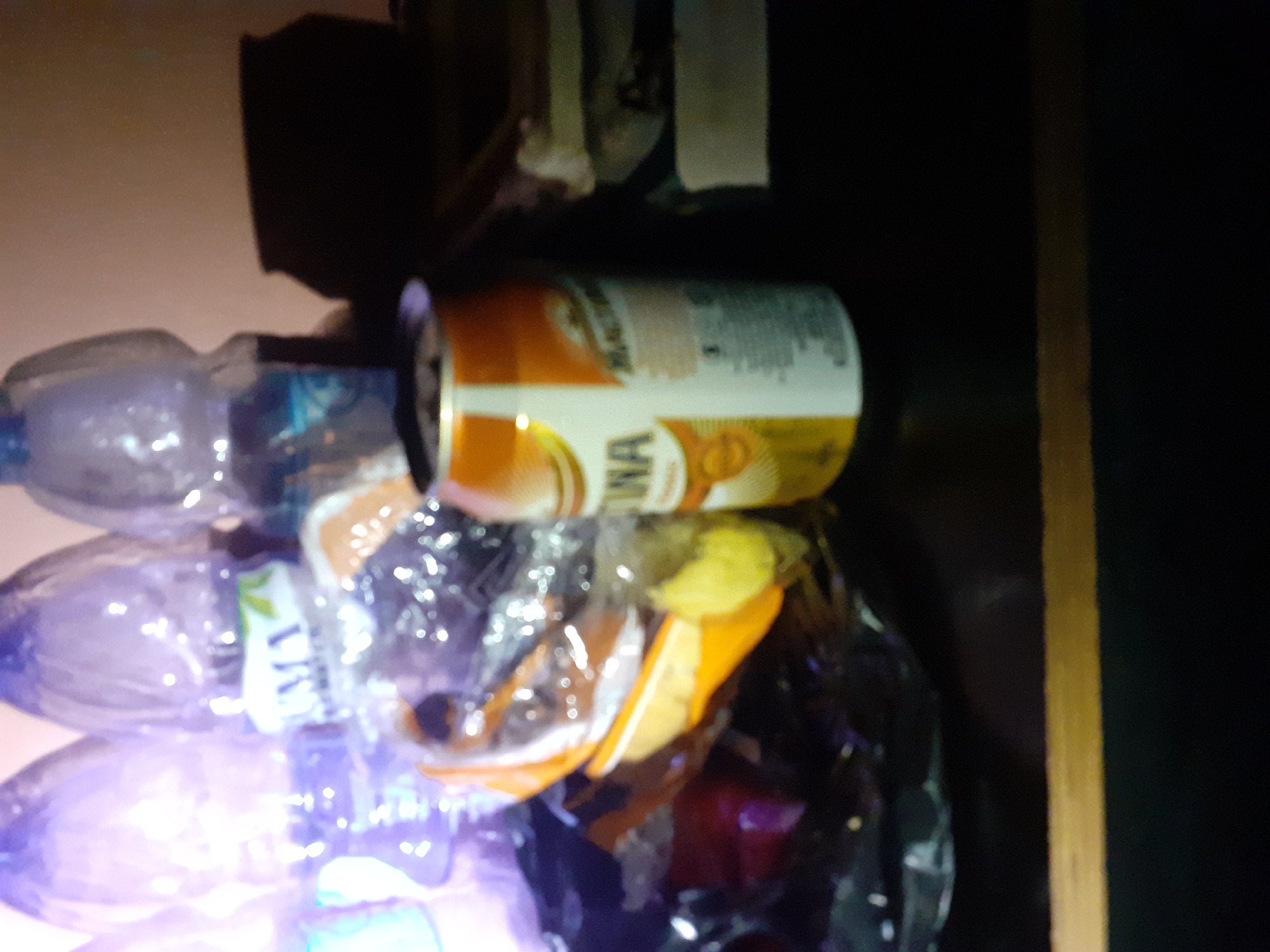 The image depicts a close-up, indoor scene with several objects on a dark surface, possibly a table. In the foreground, a partially obscured orange and white can is visible, with visible text that appears to be in a Latin-based script. To the left of the can are several clear plastic bottles, some filled with liquid and one with a white and green label. Behind the can, to the right, are stacked items that resemble books or magazines, with white and gray covers. The background is a plain, light-colored wall. The lighting is dim, casting shadows, particularly a large dark shadow to the right of the can. The overall impression is of a casual arrangement of drinks and reading material. There are no people or animals visible in the image. The location within the city of Jalingo, Nigeria, cannot be determined from the visual information provided in the image.