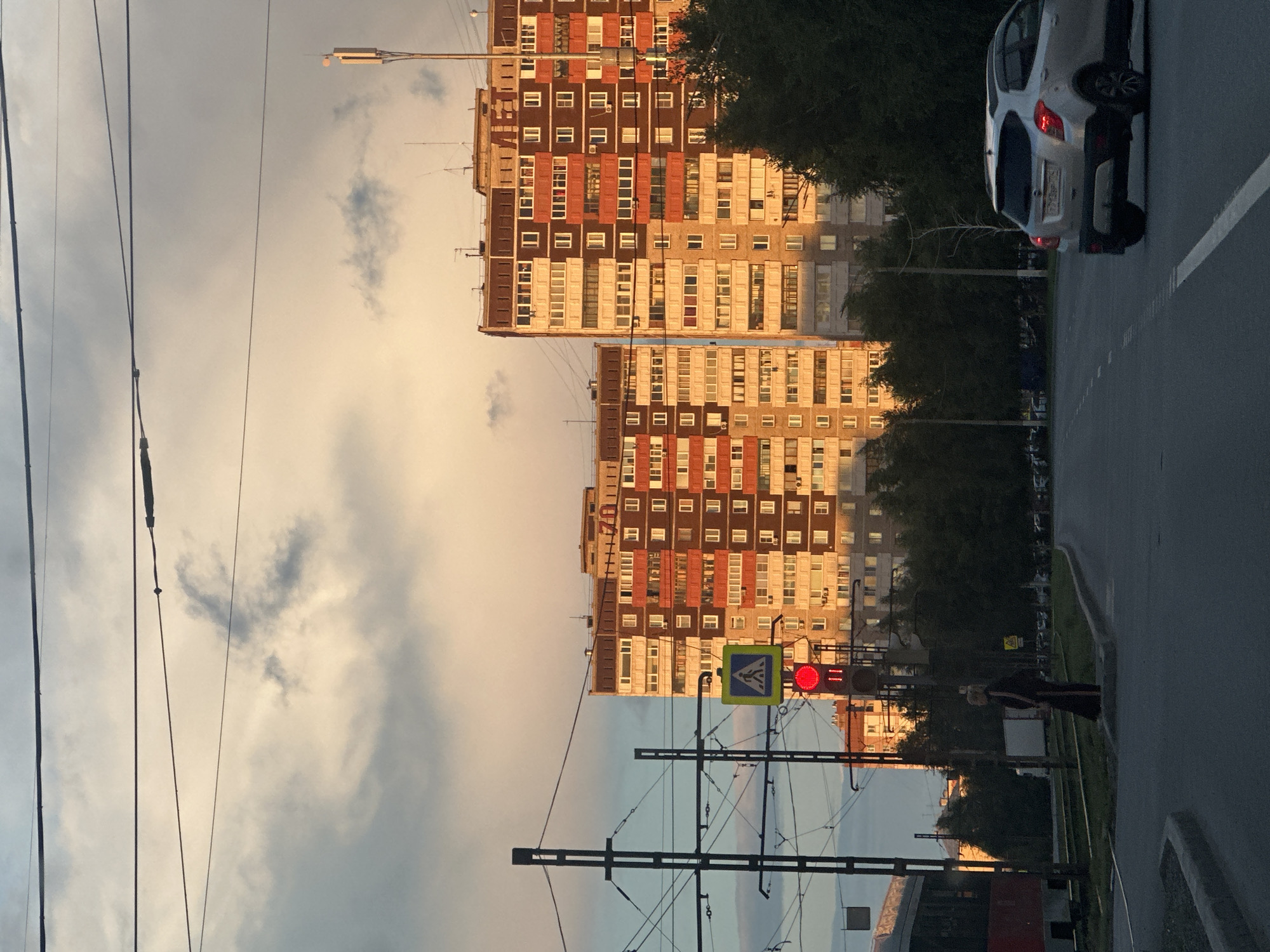 This media file, taken in Magnitogorsk, Russia, captures an urban street scene at dusk. The image is oriented vertically, with the sky and buildings dominating the frame.
The primary subjects are two large, multi-story apartment buildings with a mix of brick and beige facades, featuring numerous windows. A white car is parked on the right side of the road, its brake lights illuminated. In the foreground, there's a complex network of overhead power lines and tram wires, indicative of an urban transportation system. A traffic light shows a red signal, and a pedestrian crossing sign is visible nearby. A person, likely waiting to cross, stands on the sidewalk to the right of the traffic light, partially obscured by shadows.
The setting is a city street with a paved road and a sidewalk. The time of day is late afternoon or early evening, as evidenced by the warm, soft light illuminating the buildings and the darkening sky with scattered clouds. The overall mood is calm and ordinary, depicting a typical urban environment as day transitions into night.
There is some visible text on one of the buildings, appearing to be the number "70" in a large font.