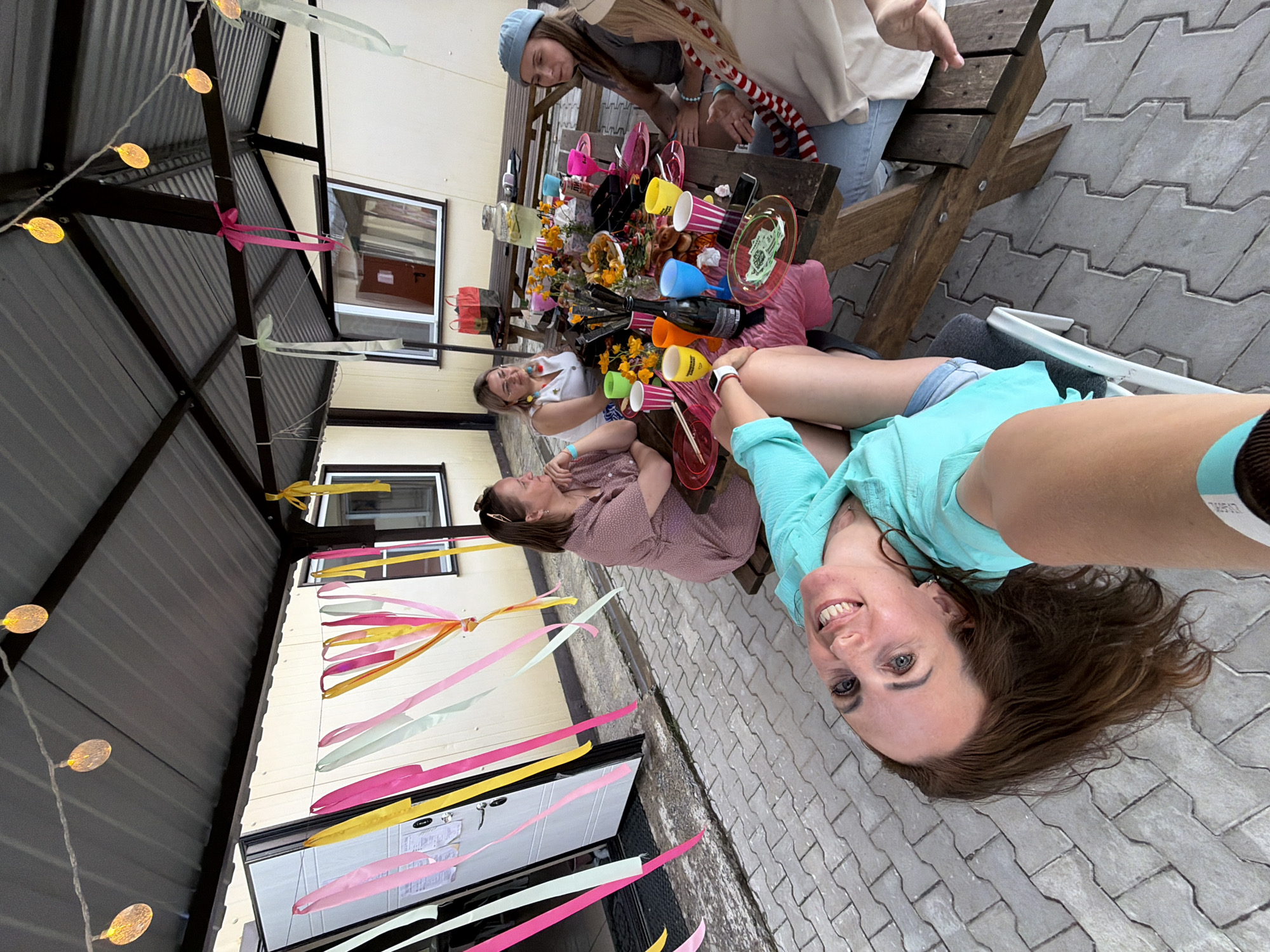 The image depicts an outdoor gathering under a covered patio. In the foreground, a woman lies on her back, looking up at the camera with a wide smile. Her hair is brown and falls around her shoulders. She is wearing a turquoise top. Behind her, a wooden picnic table is laden with food, drinks, and colorful plastic cups and plates. Four other women are seated at the table, engaged in conversation. They are dressed casually. The environment is decorated with colorful streamers and string lights, suggesting a festive occasion. The ground is paved with interlocking gray bricks. In the background, there is a building with a white exterior and windows. The scene appears to be in a residential or communal outdoor area. The image does not provide enough specific visual cues to determine its exact location within Birobidzhan, Russia.