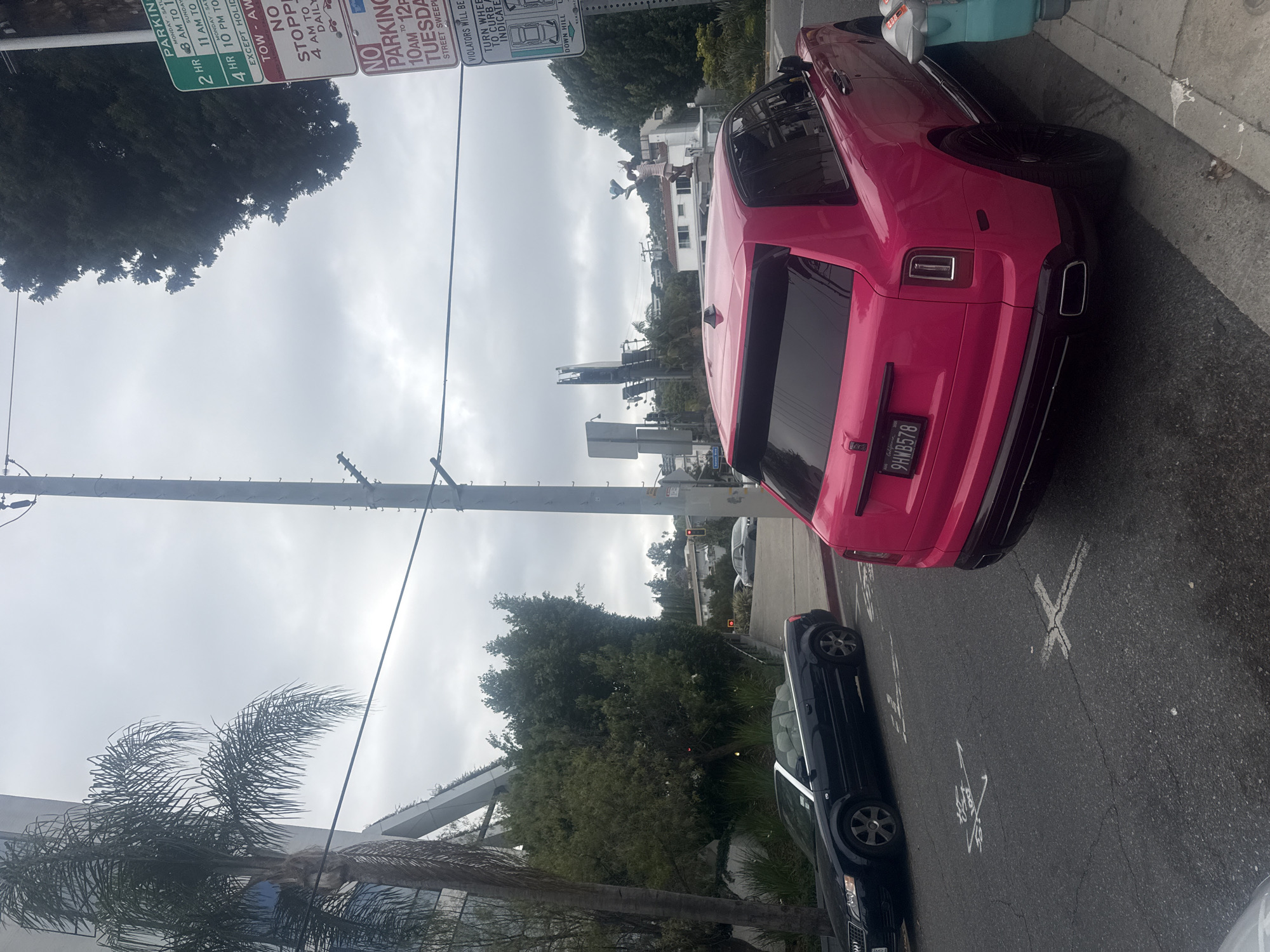The image depicts a pink Rolls-Royce parked on a street in West Hollywood, United States of America. A black sedan is parked in front of it. There are parking signs nearby. The vehicle's license plate reads 9HWB578. Street markings indicate the direction of parking. The weather appears overcast. A palm tree is visible on the left. In the background, buildings and trees are present. Electrical wires stretch across the sky.