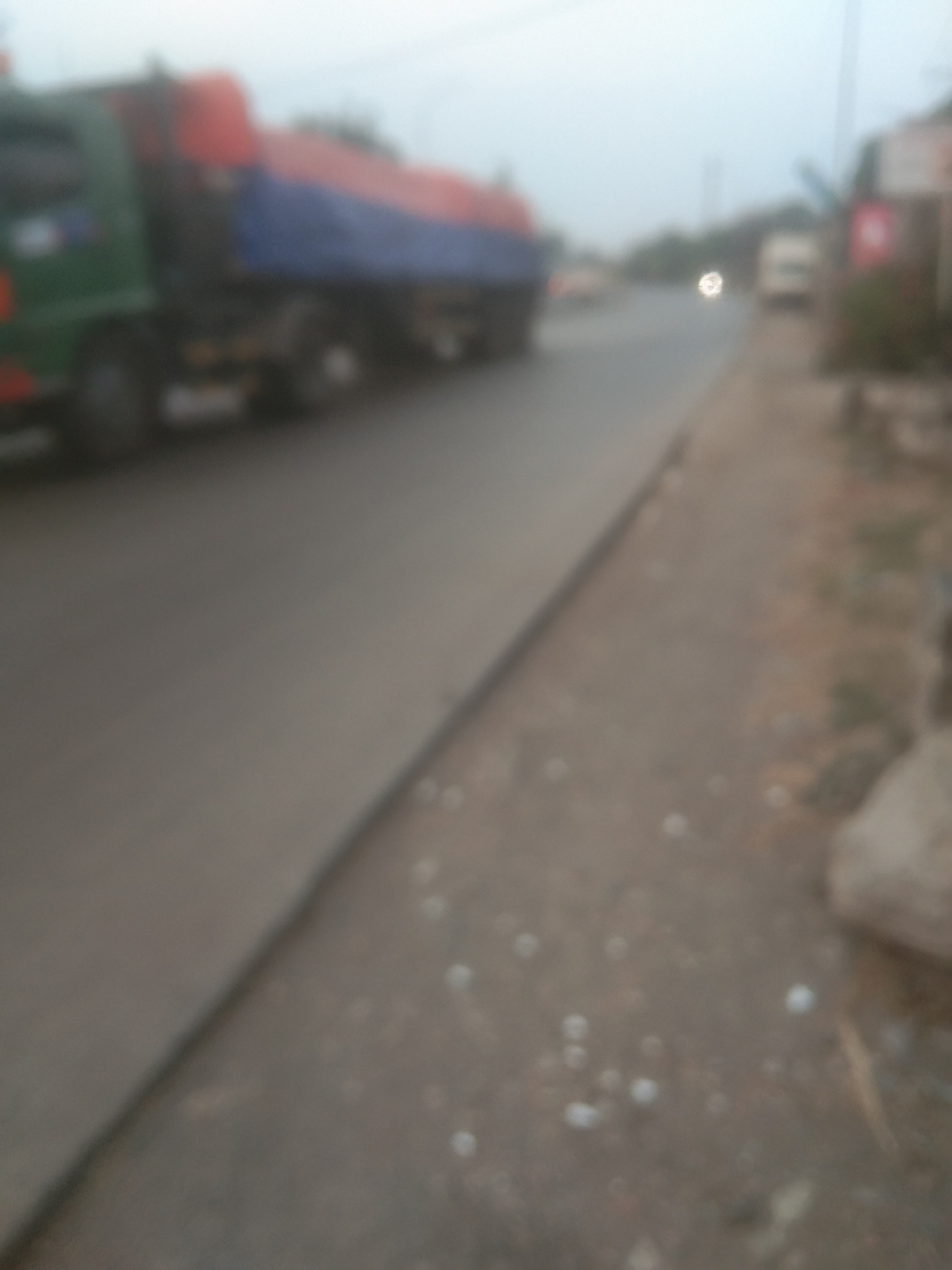 The image depicts a street scene in Subang, Indonesia, characterized by a blurred and hazy atmosphere, suggesting either poor lighting conditions or an intentionally softened focus. In the foreground, a large truck with a green cab and a red and blue cargo container is visible, positioned on the left side of the road. The truck appears to be in motion or parked. The road itself is paved and stretches into the distance. To the right of the road, a dusty embankment is present, with scattered debris. Further down the road, other vehicles, possibly vans and cars, are faintly discernible. Buildings, possibly shops or residences, are visible on the right side of the street, also out of focus. The overall impression is a snapshot of everyday urban life in Subang, with a focus on transportation and the surrounding environment.