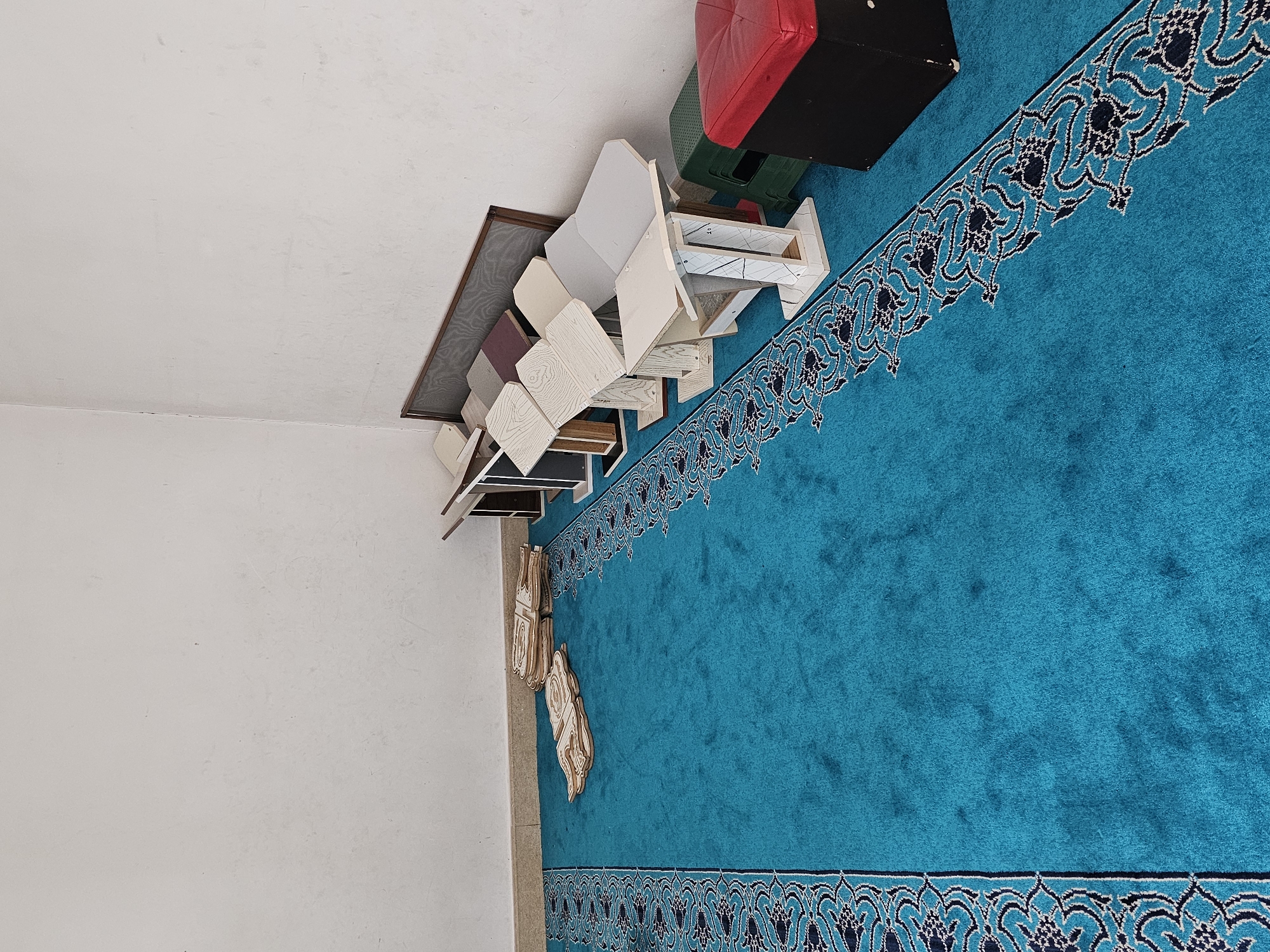 The image depicts an indoor scene with a bright turquoise carpet featuring a detailed black and white border pattern. In the background, a white wall meets a light-colored floor at the base. Several wooden lecterns of varying heights and colors, mostly white and light wood, are arranged against the wall. Some of the lecterns have a patterned grey fabric backing. To the right, a green plastic stool and a black cube-shaped ottoman with a red upholstered top are visible. In the foreground, on the left side of the carpet, several ornate wooden cutouts are stacked. The overall impression is one of a storage area or a preparation space for religious items, possibly within a mosque or a similar place of worship. The scene is static and there are no people present. The location is identified as the city of Özgön, Kyrgyzstan.