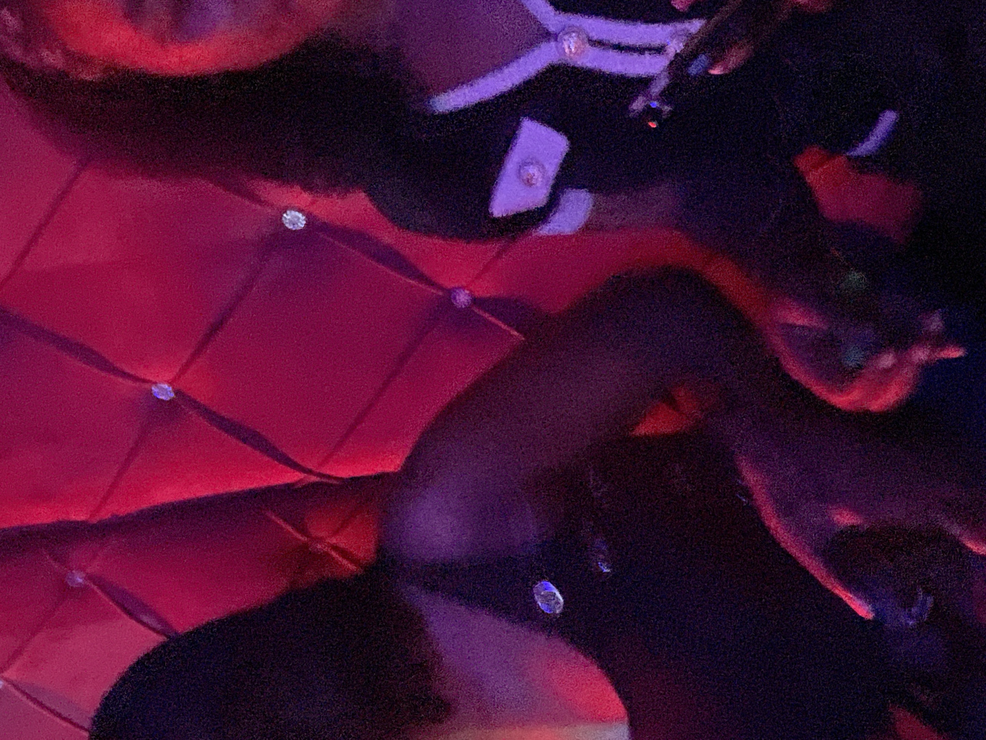The image shows two women in a dimly lit, richly decorated setting with a plush red tufted background. The woman on the left is wearing a dark, possibly black, sleeveless top. A circular, reflective embellishment is visible on her chest. The woman on the right is wearing a dark jacket with white trim and decorative buttons. She is holding a phone in her right hand. The lighting is primarily red and purple, creating a vibrant and intimate atmosphere. The scene appears to be indoors, likely in a lounge or entertainment venue in Durimi, Nigeria, suggested by the available context.