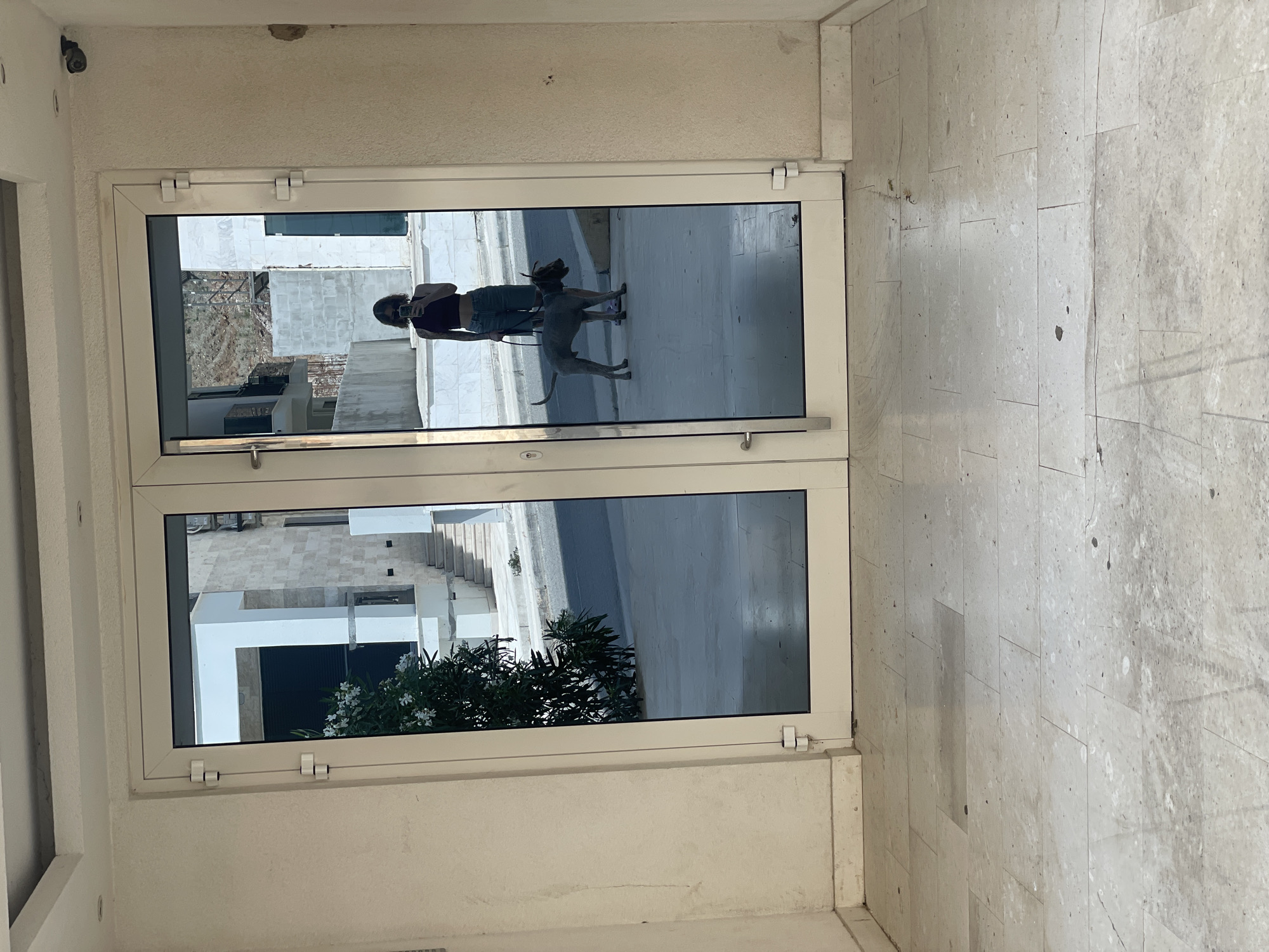 The image shows an interior view of a building, likely in Boreti, Montenegro, as suggested by the reflected architecture. The focal point is a set of closed, double-glazed doors with white frames. The doors reflect an outdoor scene: a woman in casual clothing is standing with a grey dog on a leash. The surrounding outdoor environment, reflected in the doors, includes light-colored, modern buildings, stairs, and some greenery. The interior shows light-colored tiled flooring and walls. A surveillance camera is visible in the upper left corner of the wall above the door.