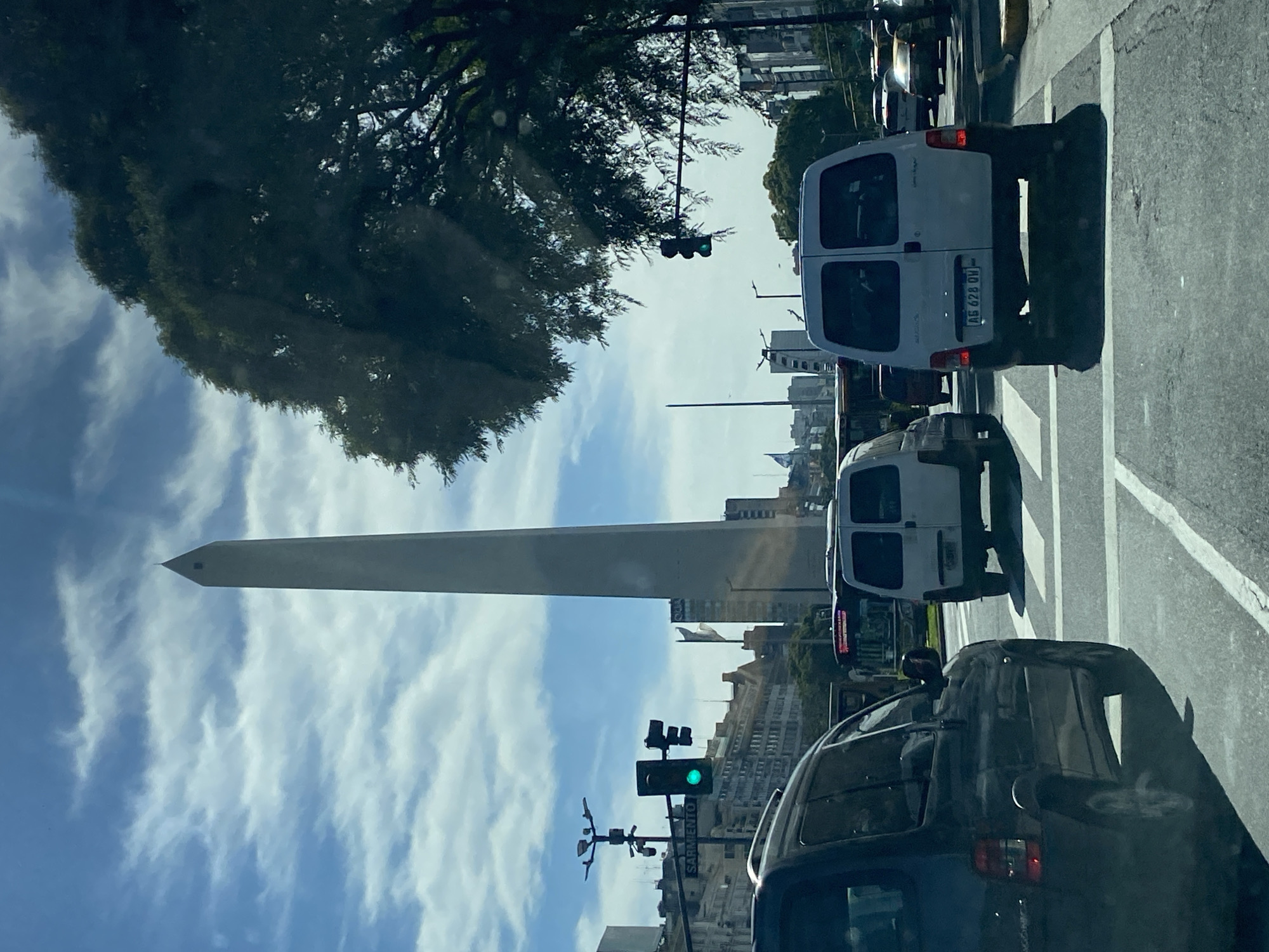 The image depicts a bustling daytime street scene in Buenos Aires, Argentina, observed from inside a vehicle. The iconic Obelisco de Buenos Aires stands prominently on the left side of the midground, a tall, slender monument piercing the bright blue sky which is dotted with scattered white clouds.
In the foreground, the dark roof and windshield of the viewer's vehicle are visible on the right, suggesting the photograph was taken from a car. The asphalt road features clear white lane dividers and crosswalk markings. Directly ahead, a white commercial van is stopped or moving slowly, its rear clearly visible. The license plate on this van reads "AB 628 OV," consistent with Argentine vehicle registration. Further down the street, another white van and other cars are visible, contributing to the urban traffic.
A traffic light, glowing green, signals movement or readiness to proceed, and a street sign below it clearly indicates "SARMIENTO," identifying a major avenue. On the left side of the street, large, leafy green trees line the sidewalk, providing shade. Beyond the immediate street level, various multi-story buildings, typical of an urban landscape, form the backdrop. The overall lighting suggests it is a clear day, likely late morning or early afternoon, with ample sunshine.