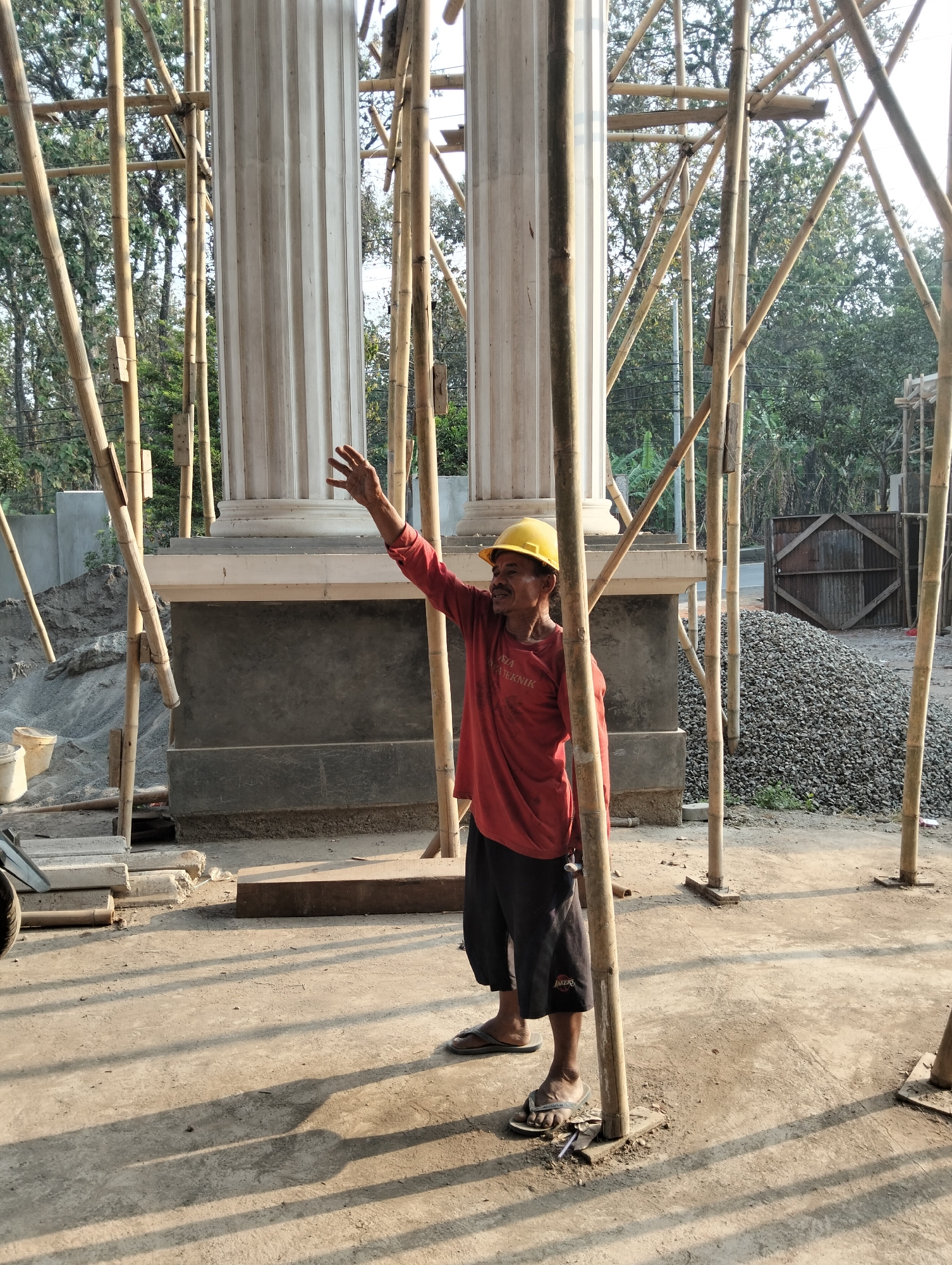 An outdoor, medium shot shows a man in a yellow hard hat and red long-sleeved shirt standing near two large, fluted columns that appear to be under construction. The columns are white and have a decorative capital. Bamboo scaffolding surrounds the columns. The man is facing left with his left arm raised and hand extended as if gesturing. He is wearing black shorts and sandals. In the background, there is a large pile of grey gravel to the right and a concrete wall and more gravel to the left. The ground is dirt and concrete, with long shadows cast by the scaffolding. The overall scene suggests a construction site in Adinuso, Indonesia.