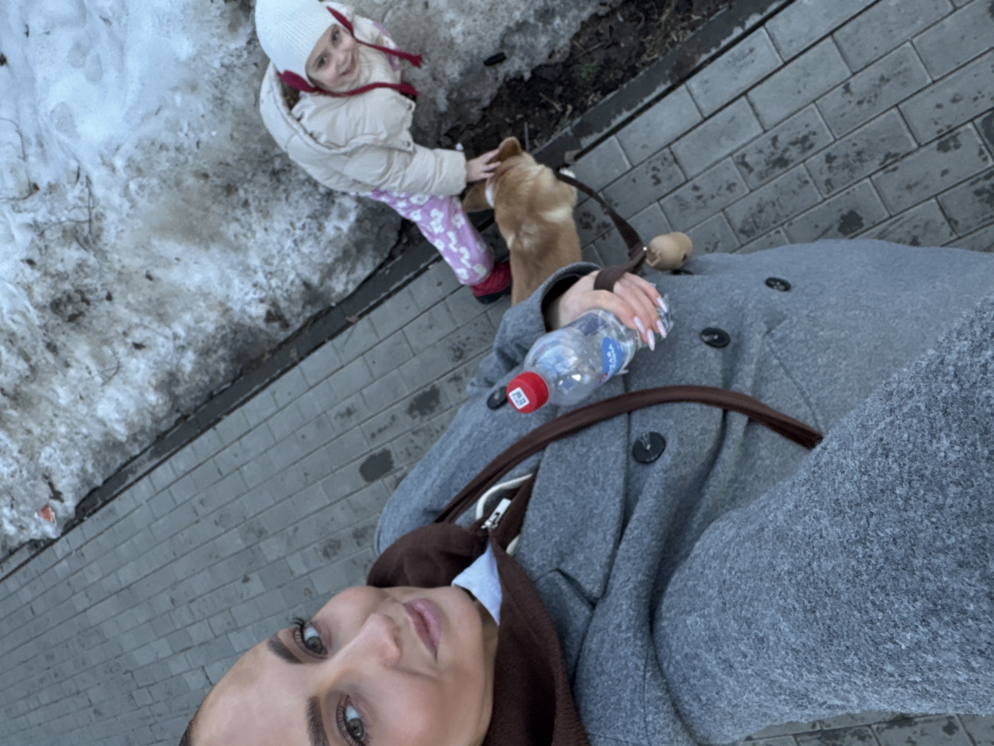 An adult female captures a high-angle selfie on a paved sidewalk in Chaikovsky, Russia. She has light-colored eyes and wears a grey overcoat with a brown hooded layer, holding a clear plastic water bottle with a red cap in her right hand. Her nails are manicured light pink. To her right, a young child, wearing a beige winter jacket, a white beanie with red ties, pink patterned pants, and red boots, smiles up at the camera while petting a light-brown dog on a leash. The dog stands near the child's feet. The environment consists of grey rectangular paver blocks on the left and a strip of dirty, melting snow mixed with dark earth and debris to the right, indicating a cold weather season.
