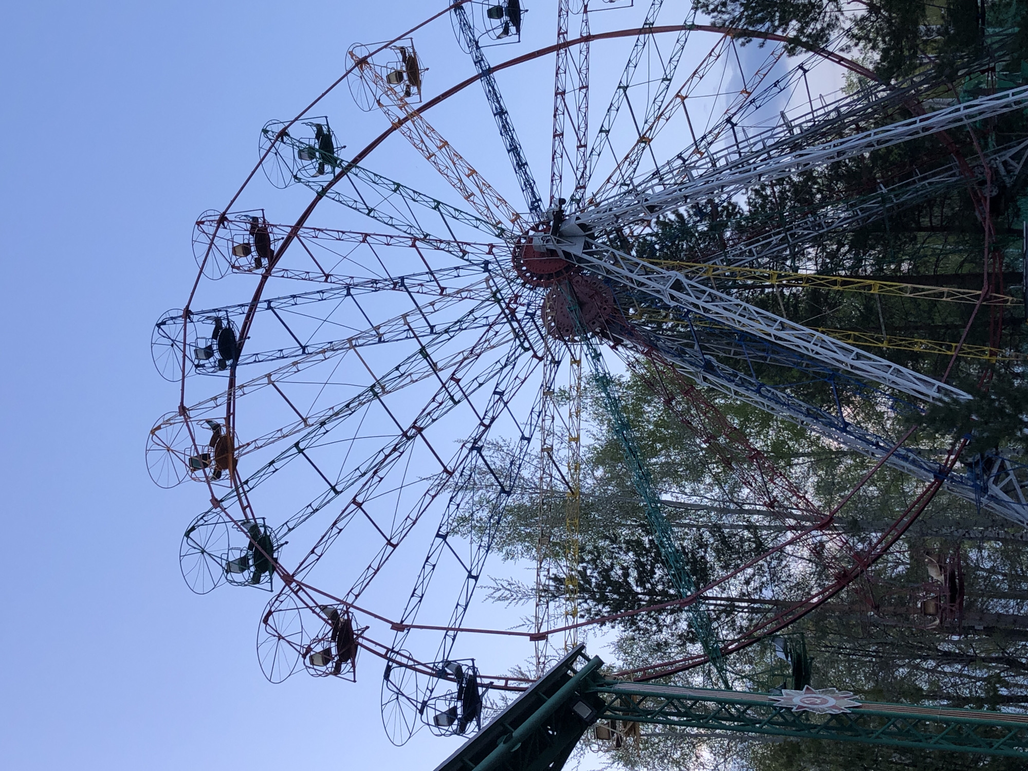 The image shows a Ferris wheel against a blue sky and trees, located in Chaikovsky, Russia. The Ferris wheel has a colorful structure with red, white, blue, yellow, and green supports. The passenger cars are open and cage-like, some appearing to be occupied. The foreground includes green metal supports and trees that partially obscure the lower portion of the Ferris wheel.
