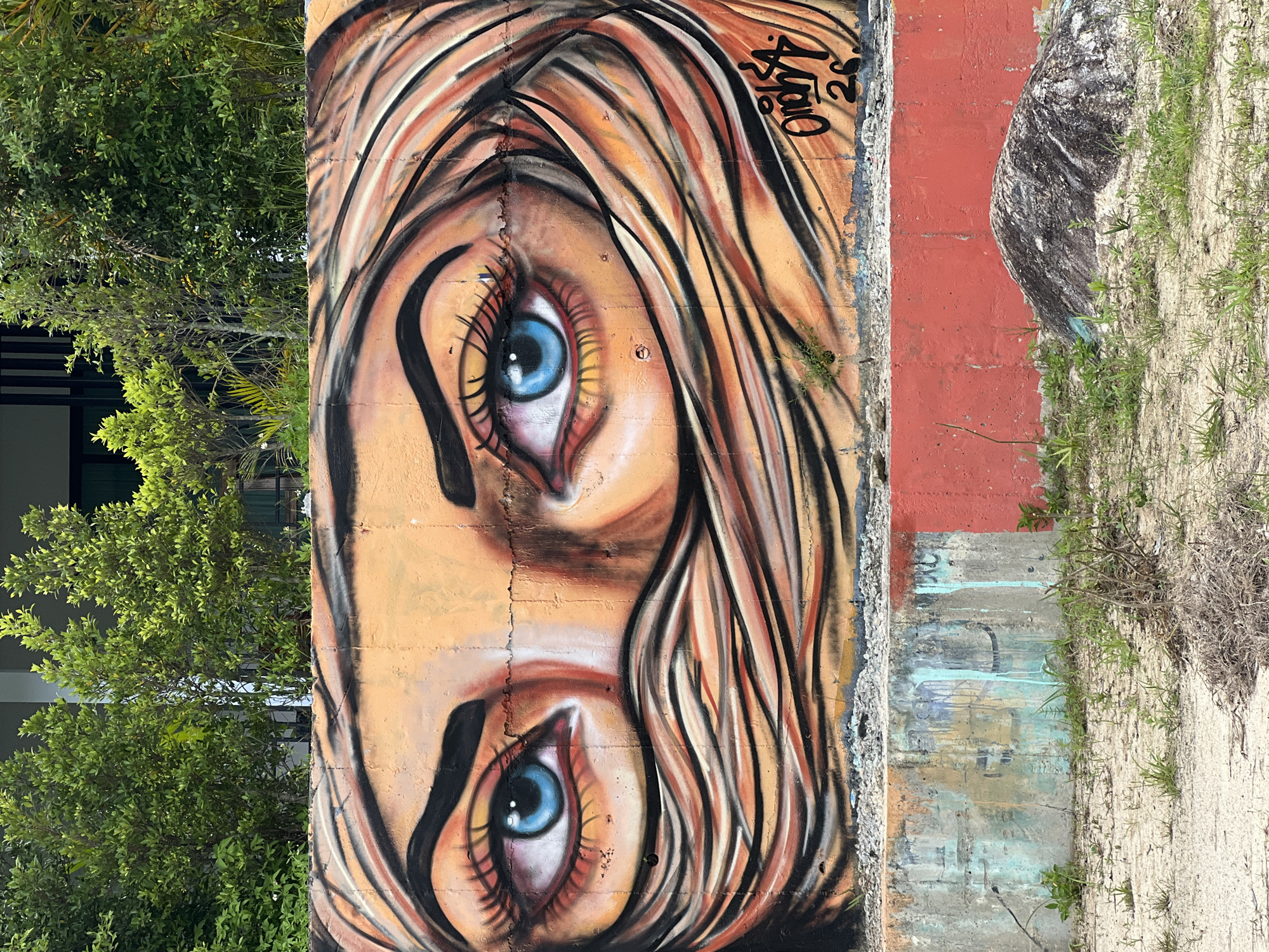 The image shows a colorful graffiti artwork of a woman's eyes on a concrete wall. The eyes are a striking blue, and the artwork depicts strands of hair or fabric flowing around her face. The wall is situated outdoors, with lush green trees and foliage visible in the background. In front of the graffiti wall, there is a lower concrete structure, partially painted in red and with some blue and yellow smudges. The ground in front of the wall is covered with sand and sparse vegetation. To the right, a large, textured rock sits on the ground. The graffiti includes a signature and a date: "OMYSA 26". The scene is likely located in a public area, possibly an urban or semi-urban environment in ตำบลเกาะพะงัน, Thailand, given the tropical vegetation and the style of construction.