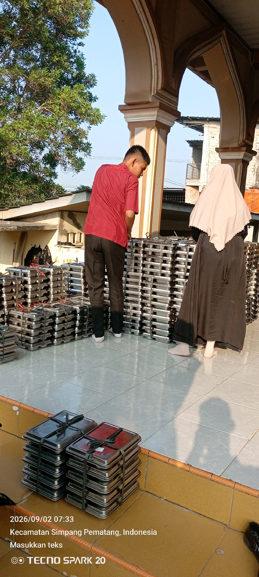The image depicts an outdoor scene in Simpang Pematang, Indonesia, likely near a building with arched architectural elements. The ground is tiled, and stacks of metal trays, possibly for catering or food service, are prominently displayed. Two individuals are visible: a person in a red shirt and black pants stands facing the trays, while another person wearing a headscarf and a dark abaya stands with their back to the camera, observing the trays. The atmosphere suggests a preparation or distribution event. The timestamp and location "2026/09/02 07:33, Kecamatan Simpang Pematang, Indonesia" are overlaid on the bottom left of the image, along with the text "Masukkan teks" and "TECNO SPARK 20".